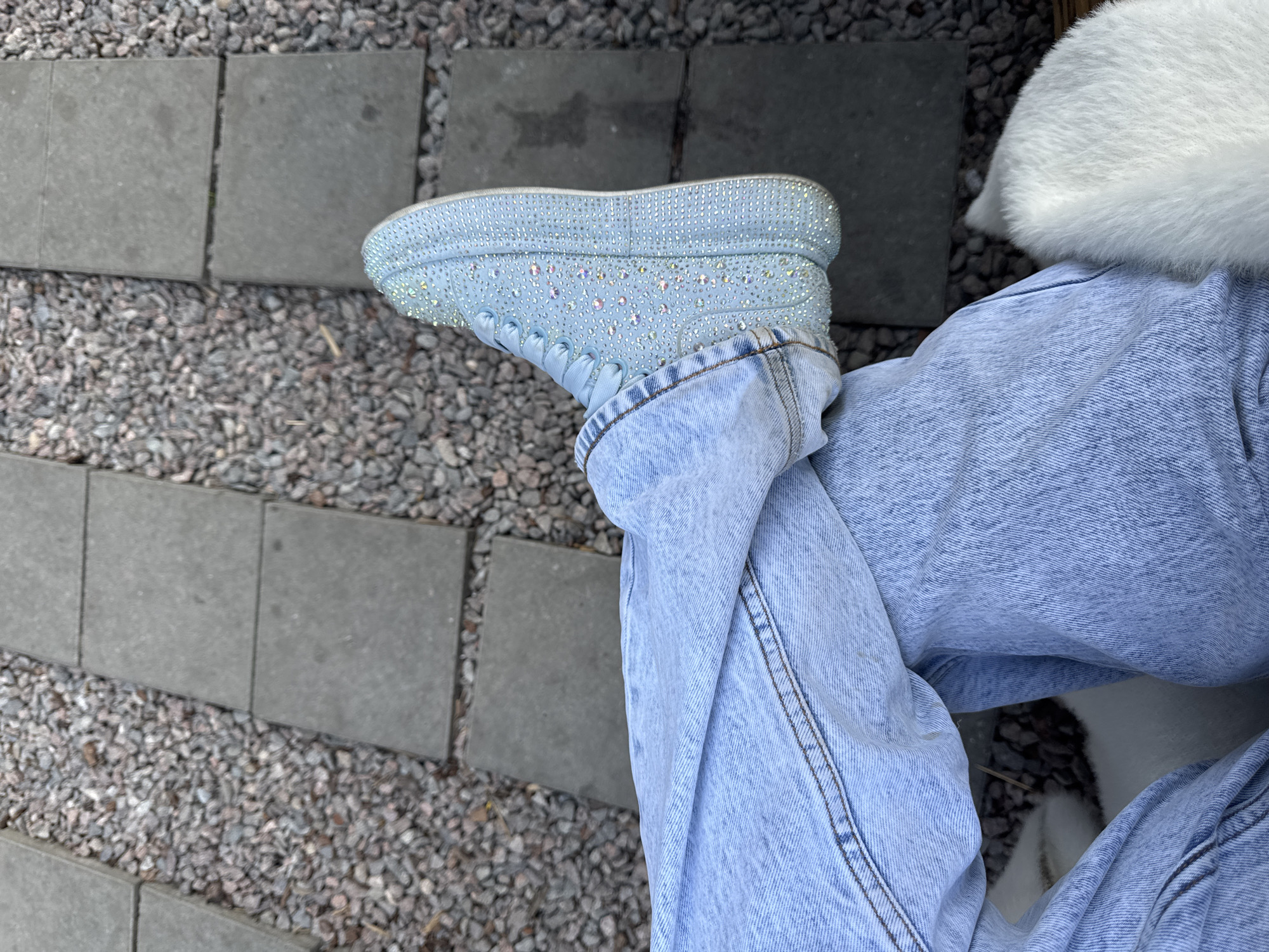 This media file is a close-up, overhead shot of a person's legs and footwear. The subjects are a pair of light blue, rhinestone-encrusted sneakers and light blue denim jeans. The setting is an outdoor area with a ground surface composed of gravel and square, gray paving stones arranged in a grid pattern.
The person's right leg is bent, with the foot of the rhinestone sneaker resting on the paving stones. The jeans are loosely fitted and appear to be acid-washed. A patch of fluffy white material, possibly belonging to a pet, is visible near the person's left leg and hip area. The lighting suggests it is daytime, with bright, diffused light, indicating possibly overcast weather or a shaded area. No text is visible in the image. The overall impression is casual and relaxed.