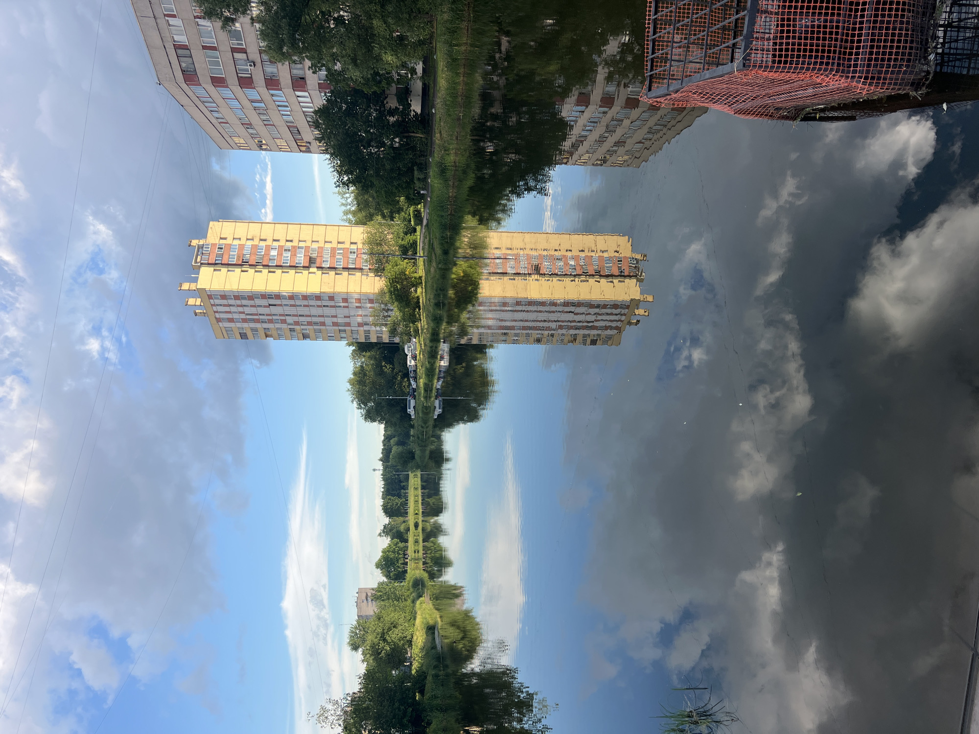 A wide-angle, full-shot, vertical-orientation, outdoor, daytime, street-level photograph captures a serene river scene in a city with two residential buildings and a reflection of the sky and buildings on the water. The sky is bright blue with scattered white clouds. The left building is tall and yellow with numerous windows and a prominent structure on its roof. The right building is lighter in color and appears to have a curved facade with balconies. Both buildings are situated behind a line of lush green trees and vegetation that grows along the riverbank. The river itself is calm, creating a clear, mirror-like reflection of the sky, clouds, buildings, and trees. In the foreground on the right, a section of a dark metal cage covered with orange netting is visible, partially submerged in the water. The overall atmosphere is peaceful and idyllic.