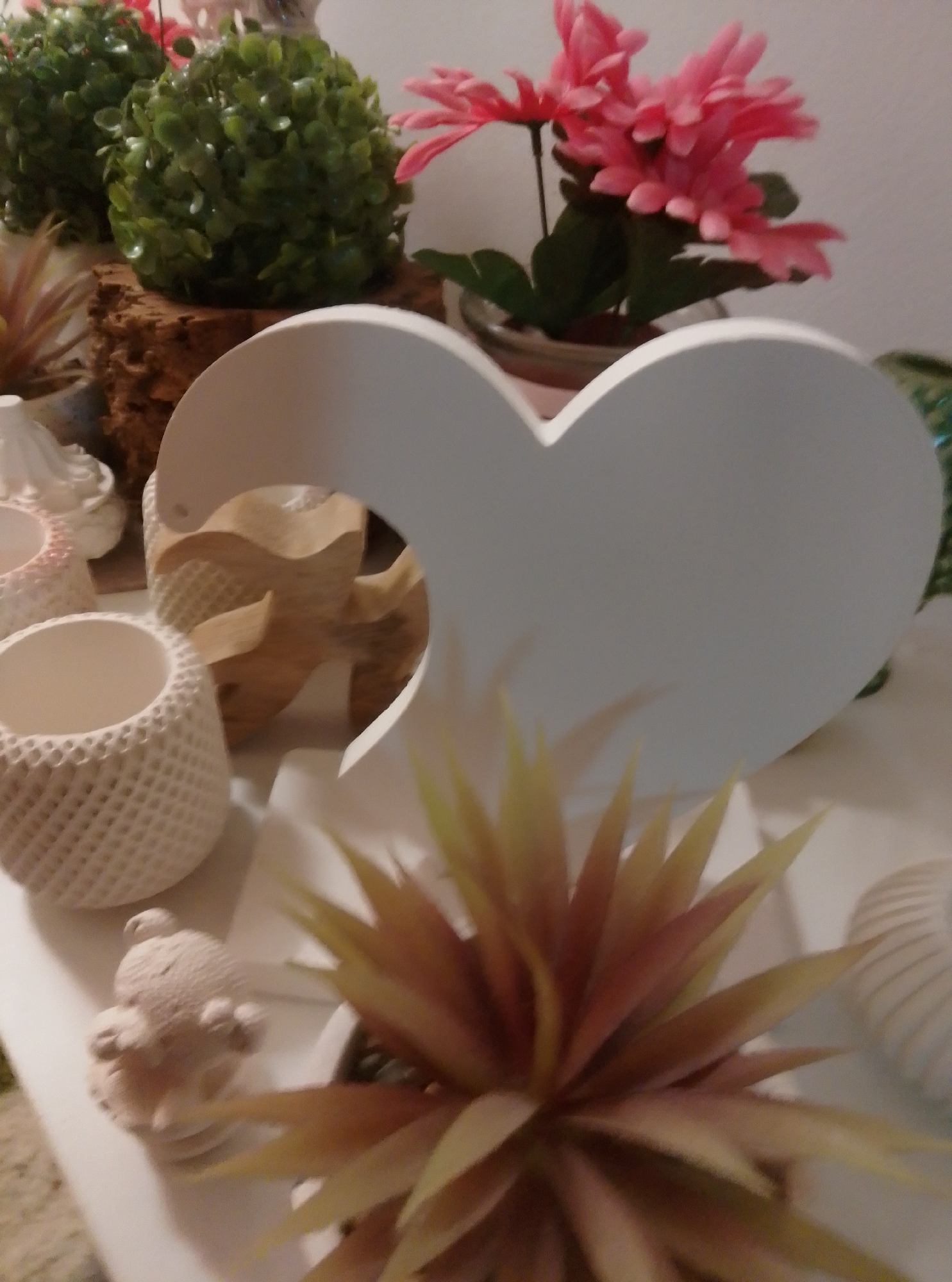 The image shows a collection of decorative items arranged on a white surface. Dominating the center is a large, white, heart-shaped cutout. To the left and behind the heart are two green spherical decorative plants. In front of the heart and to the lower right, a spiky, reddish-yellow artificial plant is visible. Other decorative items include a small, light-colored bear figurine, a textured cylindrical vase, and what appears to be a wooden starfish visible through the heart cutout. In the background, pink artificial flowers are seen. The scene is indoors, likely a home or shop display, and there is no indication of a specific location in the city of Wedel, Germany.