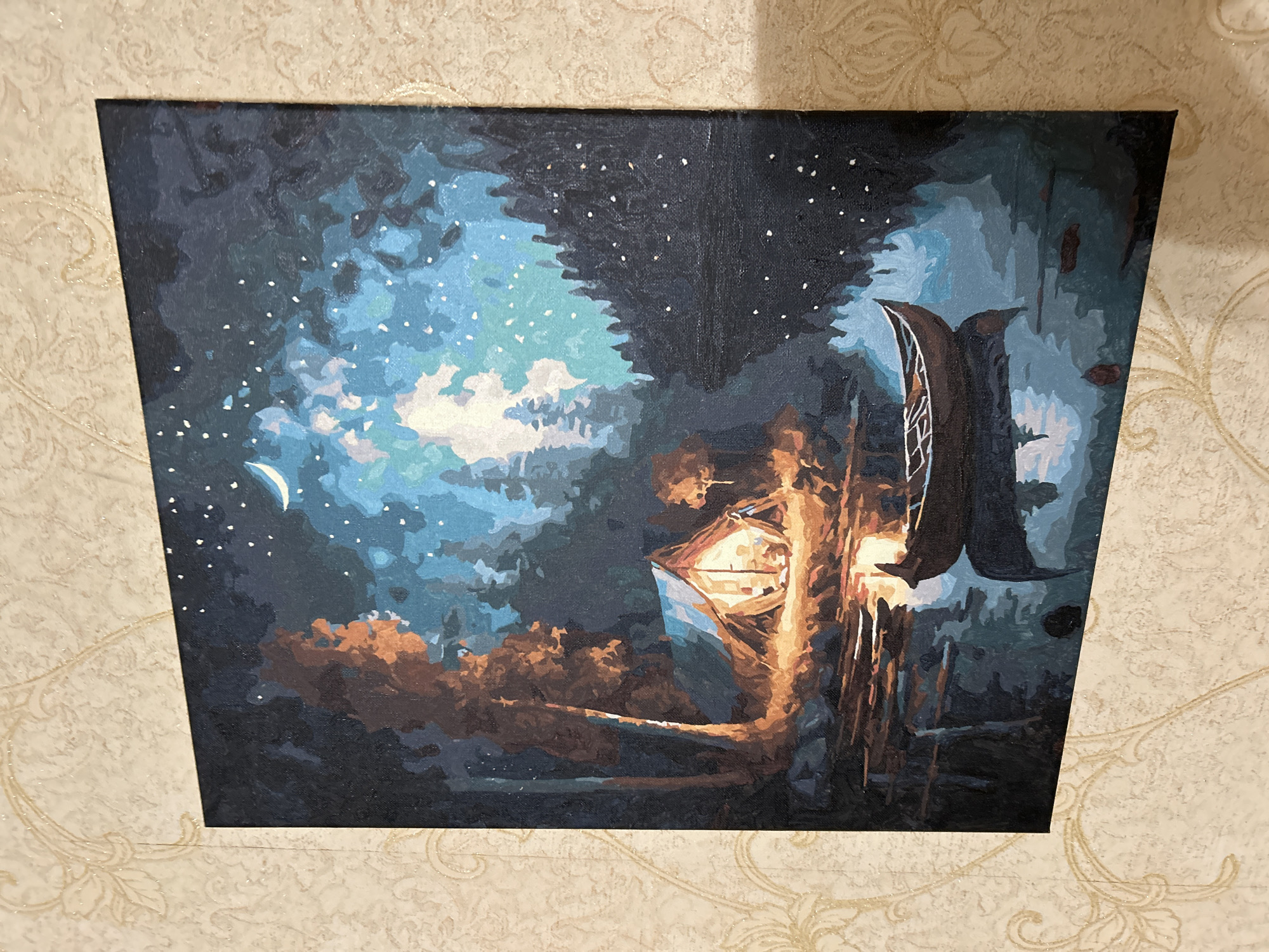 The image shows a rectangular canvas painting depicting a night scene with a predominantly dark blue and brown color palette. A crescent moon is visible in the upper left corner, accompanied by numerous small white dots suggesting stars against a dark sky. Patches of light blue and white suggest clouds or a moonlit sky. In the foreground, there is a structure, possibly a stylized building or treehouse, with visible light emanating from within. The structure is depicted with browns, yellows, and blues. The surrounding environment seems to be a stylized forest with tree silhouettes and foliage represented through dark brown and blue strokes. The bottom of the canvas is a dark, nearly black area suggesting ground or water. There is no indication of the location being in Khimki, Russia, or any other specific location. No people, specific subjects, actions, or interactions are directly visible.