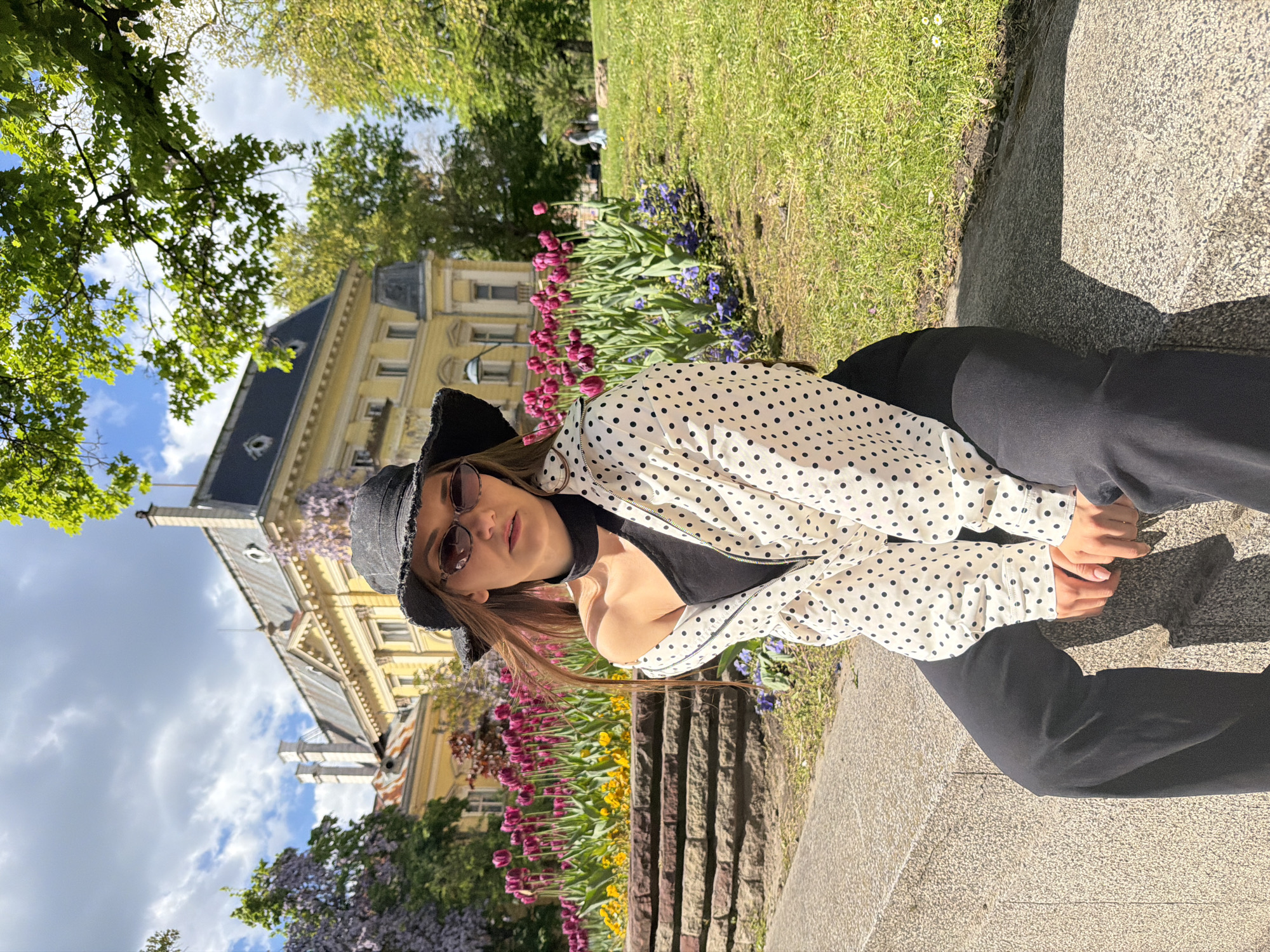 A woman is sitting outside in Sofia, Bulgaria, next to flowerbeds and a building. She is wearing a black hat, sunglasses, a black top, and a white and black polka dot jacket. She is also wearing black pants. The flowerbeds contain purple and yellow flowers. The building is yellow with a dark roof. The sky is visible with blue sky and white clouds. There are trees.