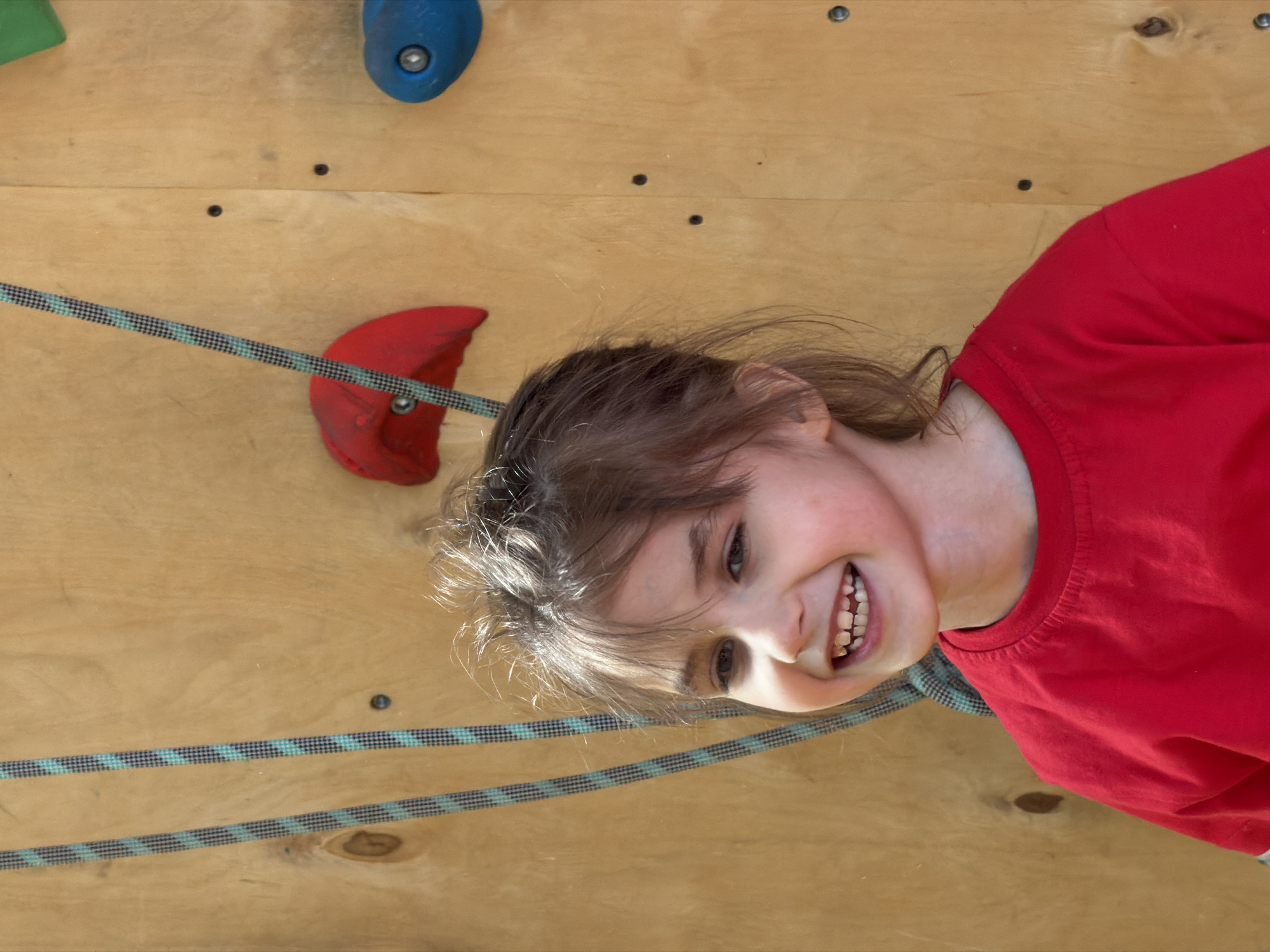 A girl in a red shirt smiles and looks towards the camera in front of a wooden climbing wall with green, blue, and red climbing holds. A black and turquoise rope spans horizontally across the wall. The location is not identifiable as Izhevsk, Russia, based solely on the provided image.