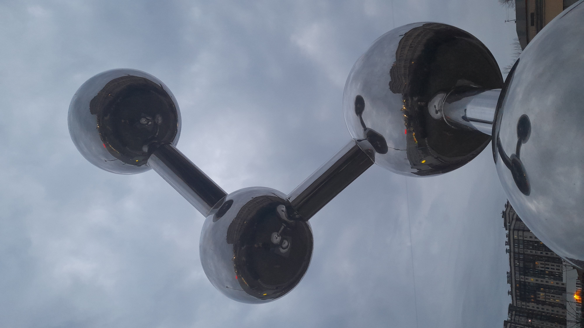 The image captures a sculpture resembling a molecule with large, reflective spheres connected by cylindrical rods. The sculpture is metallic and reflects the surrounding buildings and sky. The sky is overcast. A tall apartment building is visible in the background. The sculpture is located in Saint Petersburg, Russia, near the Novaya Gollandiya island.
