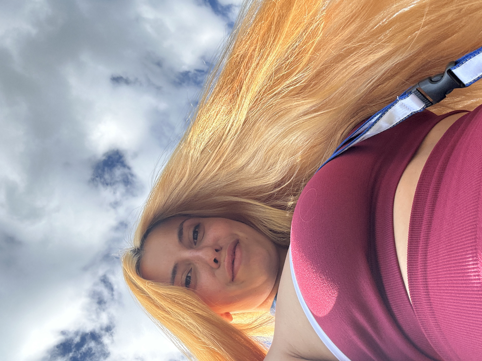 A selfie taken from a low angle depicts a person with long, straight, blonde hair, light skin, and wearing a burgundy garment with a white stripe. The person's face is visible with their head tilted, and they are looking at the camera. Above and behind the person is a blue sky dotted with white clouds. A strap with a black plastic buckle is partially visible near the top right of the frame. The location cannot be determined to be Heinebach, Germany from the provided image.