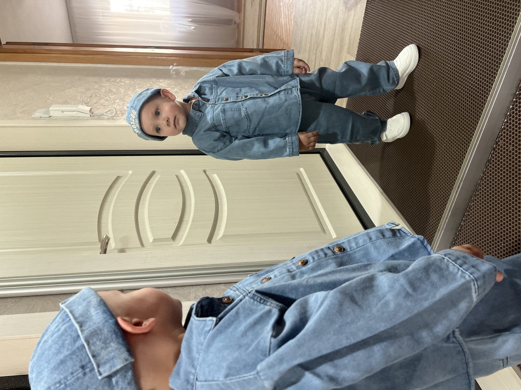 A young child dressed in a light blue denim outfit including a matching cap and white sneakers is the primary subject of the image. The child stands near a light-colored door with a silver handle. To the right of the child, a brown honeycomb-patterned floor mat is visible. The scene appears to be indoors, possibly within a home in Марковский, Russia. An additional portion of the child's outfit is visible in the lower foreground.
