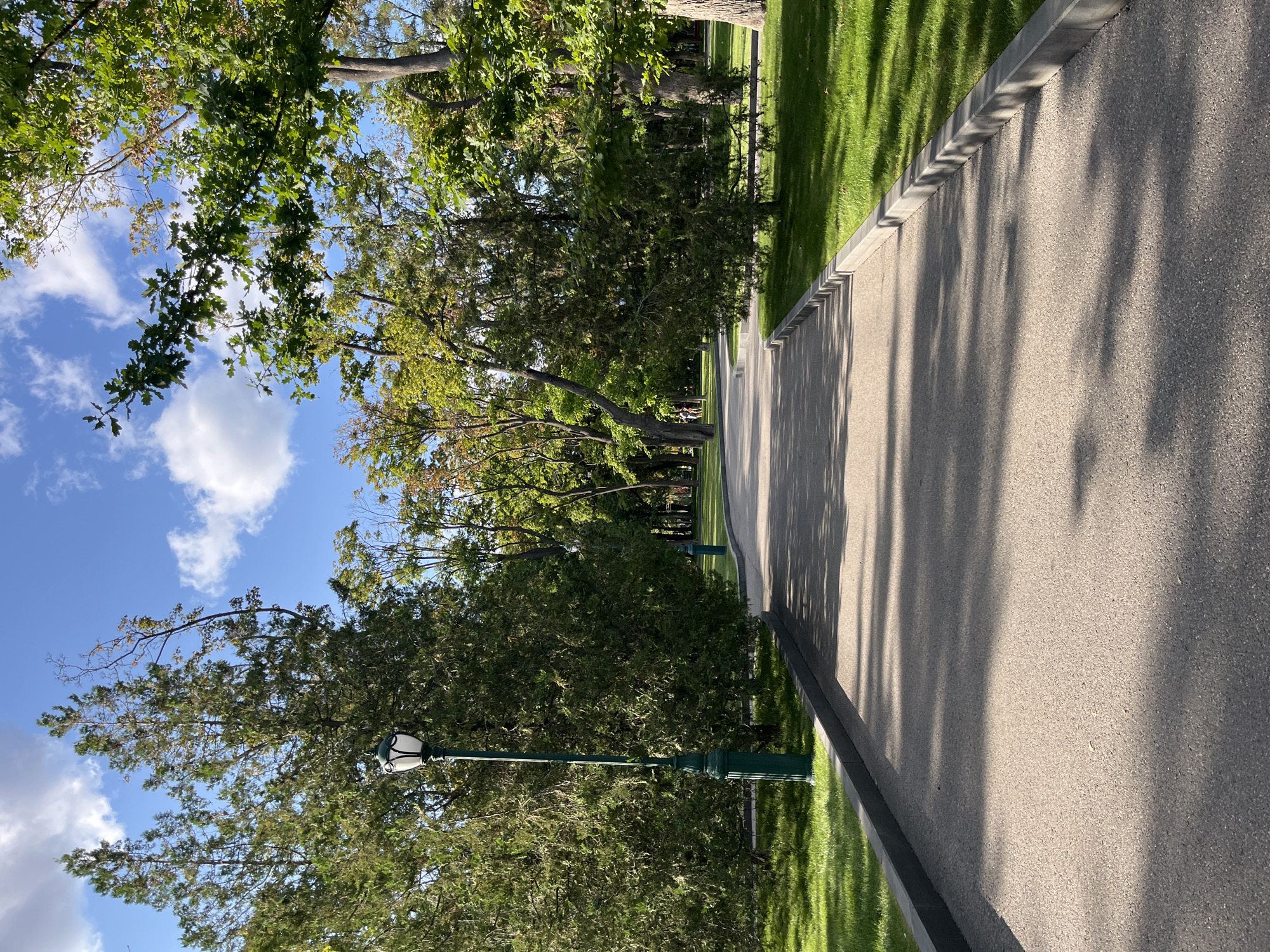 A paved pathway curves through a park in Kharkiv, Ukraine, flanked by manicured green lawns and tall, mature trees. The pathway is a light grey asphalt, bordered by dark grey concrete curbs. Sunlight casts dappled shadows from the leaves of the trees onto the pathway. On the left side of the pathway, a tall, dark green lamppost with a white lantern is visible. In the distance, the pathway continues, and several trees with varying shades of green and some autumnal yellow foliage are present. The sky is blue with scattered white, fluffy clouds. There are no people clearly visible in the image. The overall scene depicts a serene and well-maintained park environment.