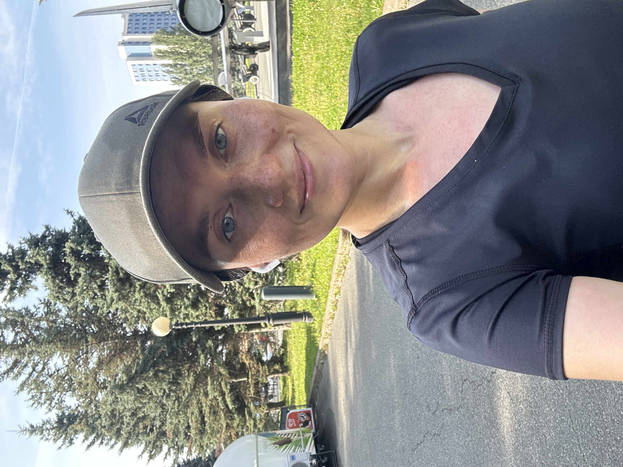 A medium-close selfie shows a woman with blue eyes and freckles smiling, wearing a dark grey t-shirt and a dark grey baseball cap with a Reebok logo. She has white wireless earbuds in her ears. She is outdoors with green trees and grass in the background. A tall, thin monument and a modern building are visible behind her. A light pole is to her left, and a white food truck with images of ice cream is parked in the distance. The ground in front of her is paved. The scene appears to be a park in Kazan, Russia, potentially near a monument and a public space.