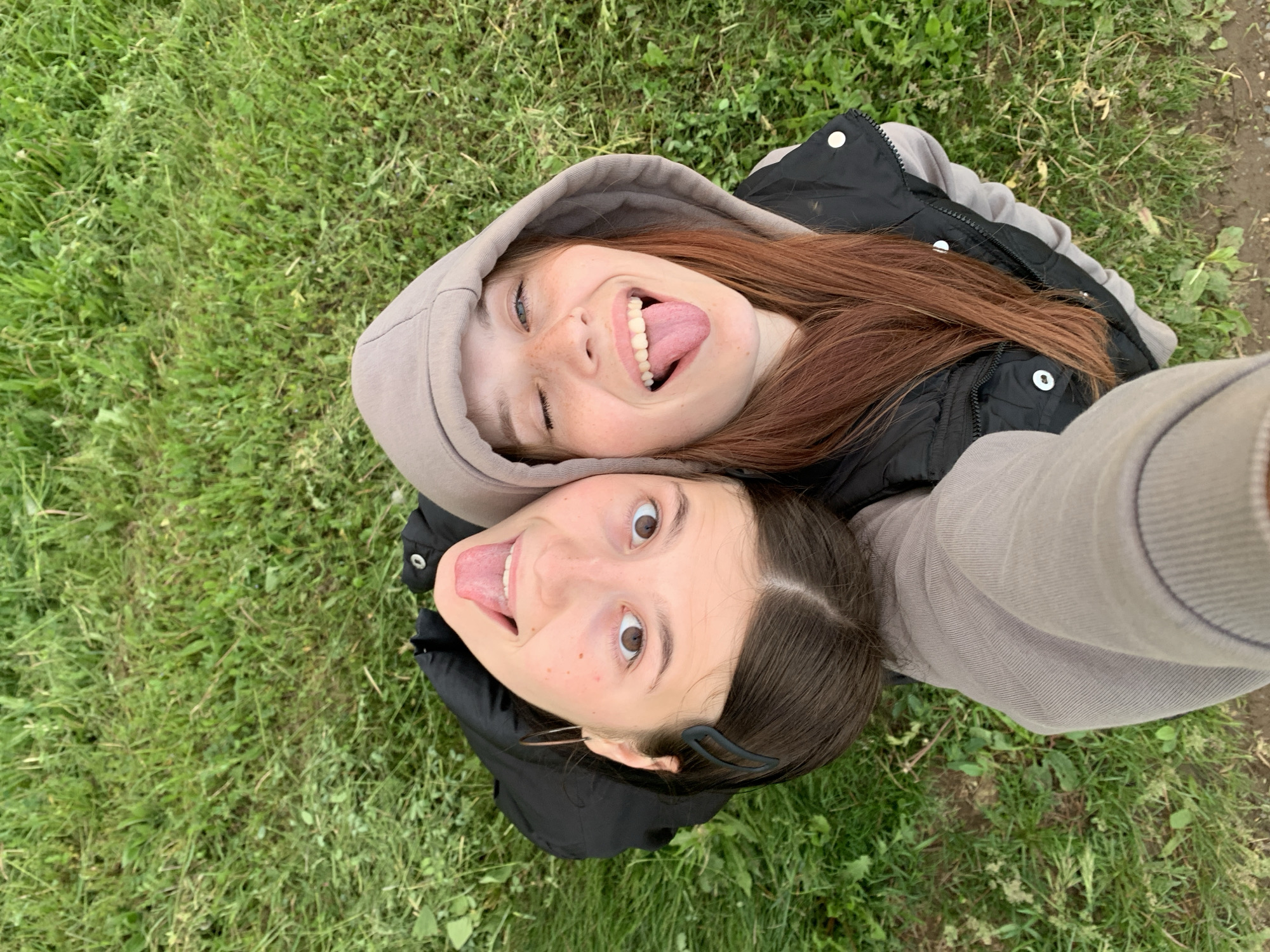 The image, taken in Фоки, Russia, is a high-angle selfie featuring two young females against a background of green grass. The subject on the left, with dark brown hair parted down the middle and secured by a black bobby pin on the left side, is positioned upside down relative to the viewer. She has wide brown eyes looking upward, and her tongue is extended. She wears a black top or vest and a silver hoop earring in her left ear. The subject on the right, with reddish-brown hair, wears a light brown hooded sweatshirt under a black sleeveless puffer vest with silver snaps. She is winking with her left eye, smiling broadly to reveal teeth, and her tongue is also extended. Both individuals exhibit playful facial expressions and have visible freckles. The arm of the subject on the right, clad in the light brown ribbed sleeve of her hoodie, is visible in the foreground, indicating she is holding the camera. The setting is outdoors with natural, even lighting.