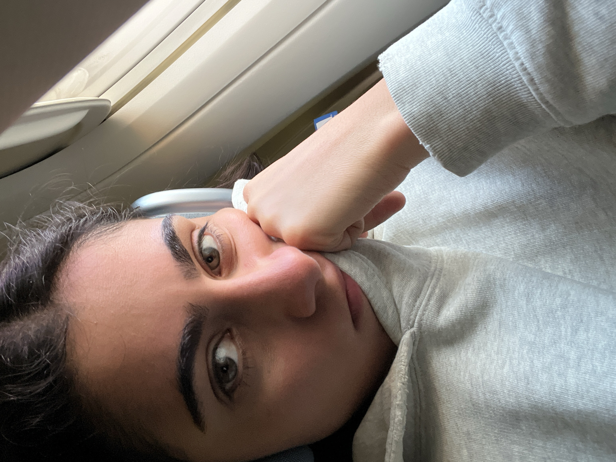 A close-up, angled selfie of a woman with dark hair and light skin is positioned in front of a window of an airplane. She is resting her face on her closed fist, which is partially covered by the sleeve of a light grey sweatshirt. The window has a white frame and multiple panels. The location is not provided in the image, so the information about Meaux, France cannot be confirmed or included.