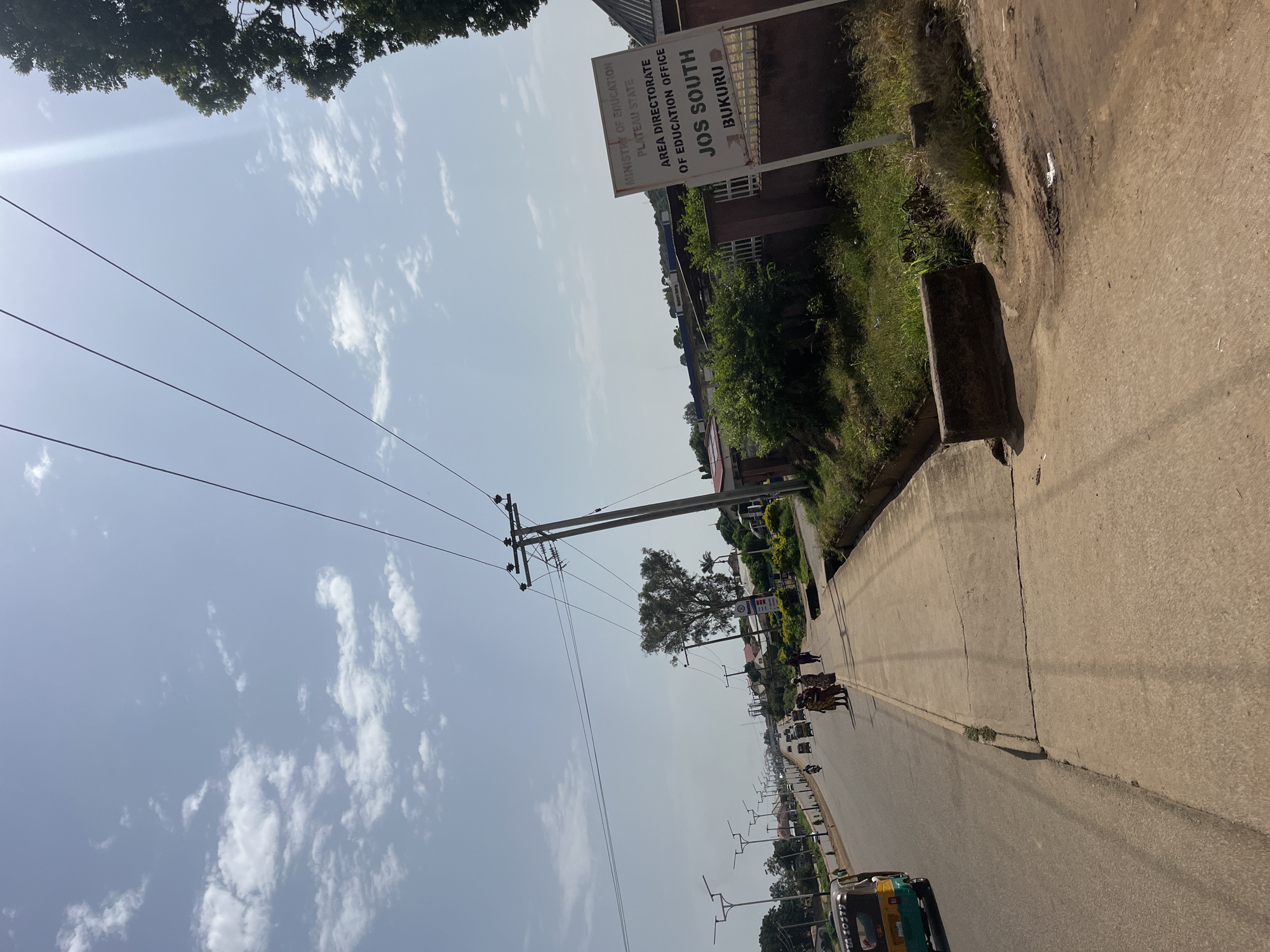 The image depicts a street scene in Bukuru, Nigeria. A paved road runs diagonally from the lower left to the upper right of the frame, with a sidewalk to its right. Several vehicles, including a three-wheeled auto-rickshaw, cars, and what appear to be minibuses or buses, are traveling on the road. A group of people, predominantly women in traditional attire, are walking on the sidewalk. On the right side of the sidewalk, there is a white signpost with black lettering that reads: "MINISTRY OF EDUCATION PLATEAU STATE AREA DIRECTORATE OF EDUCATION OFFICE JOS SOUTH BUKURU". Beyond the sidewalk and signpost, there are buildings, trees, and lush green vegetation. Overhead, power lines stretch across the sky, and there are scattered white clouds against a bright blue sky, with the sun's glare visible in the upper right corner. The overall environment appears to be a moderately busy urban street on a clear day.