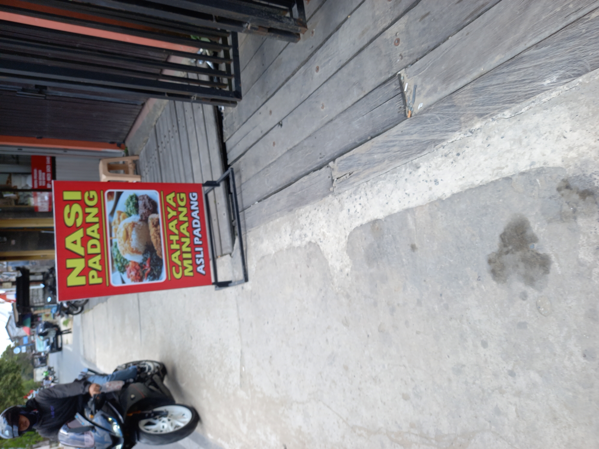 A person wearing a black helmet and black jacket rides a black sport motorcycle on a concrete sidewalk. A red and yellow sign advertising "NASI PADANG CAHAYA MINANG ASLI PADANG" is displayed on a stand to the right of the motorcycle. The sign includes an image of nasi padang. The street scene includes buildings, other motorcycles, and a wooden walkway to the right of the sign. The image appears to have been taken in Samarinda, Indonesia, based on the signage.