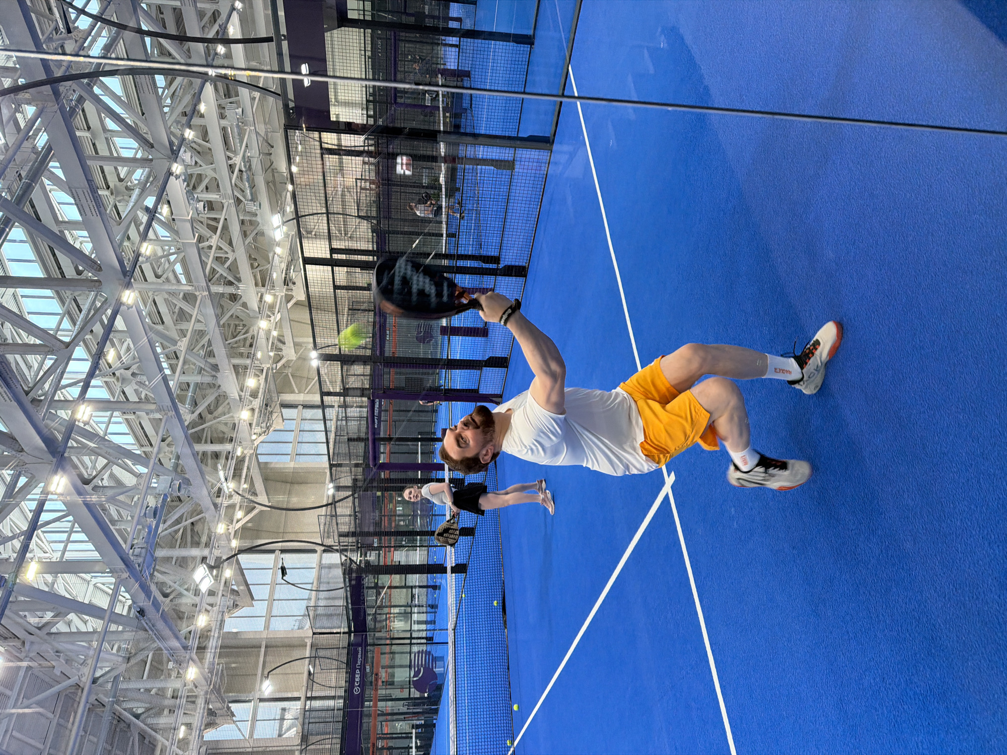 The image shows a man playing paddle tennis in Moscow, Russia. The man is hitting a ball with a paddle on a blue indoor court. He is wearing a white t-shirt and yellow shorts. There is another person on the court behind him. The court is surrounded by netting and glass walls. The roof of the building has a complex network of beams.