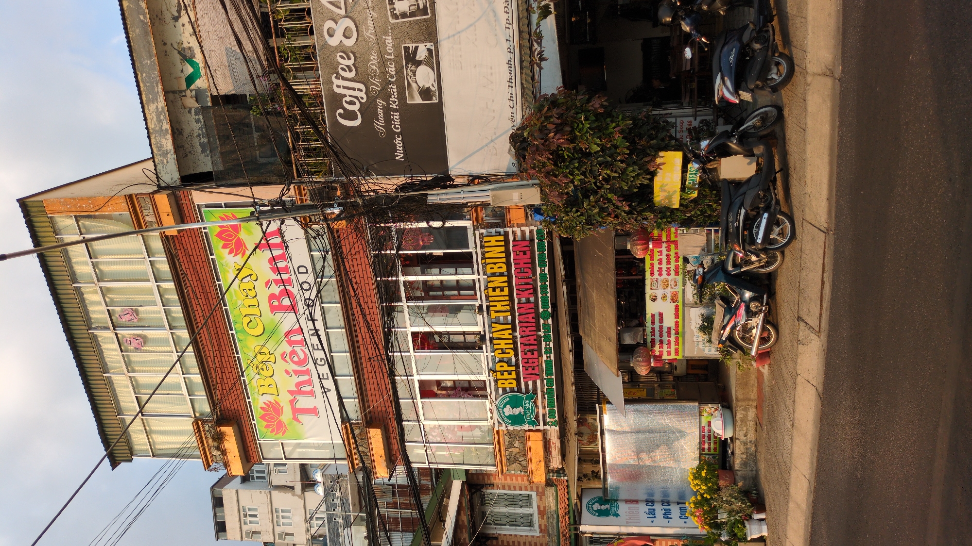 The image shows a commercial street in Da Lat, Vietnam, featuring a two-story building. The building houses several businesses: "Bếp Chay Thiên Bình VEGENFOOD," a vegetarian restaurant. A coffee shop, "Coffee 84," is also located above the restaurant. "Bếp Chay Thiên Bình VEGETARIAN KITCHEN" has a sign in both Vietnamese and English. Motorbikes are parked along the curb in front of the building. There are multiple signs, including menu boards and promotional materials for the restaurant. The street is congested with utility wires. The address "Nguyễn Chí Thanh, p. 1, Tp. Đà" is visible. There are plants and flowers along the sidewalk.