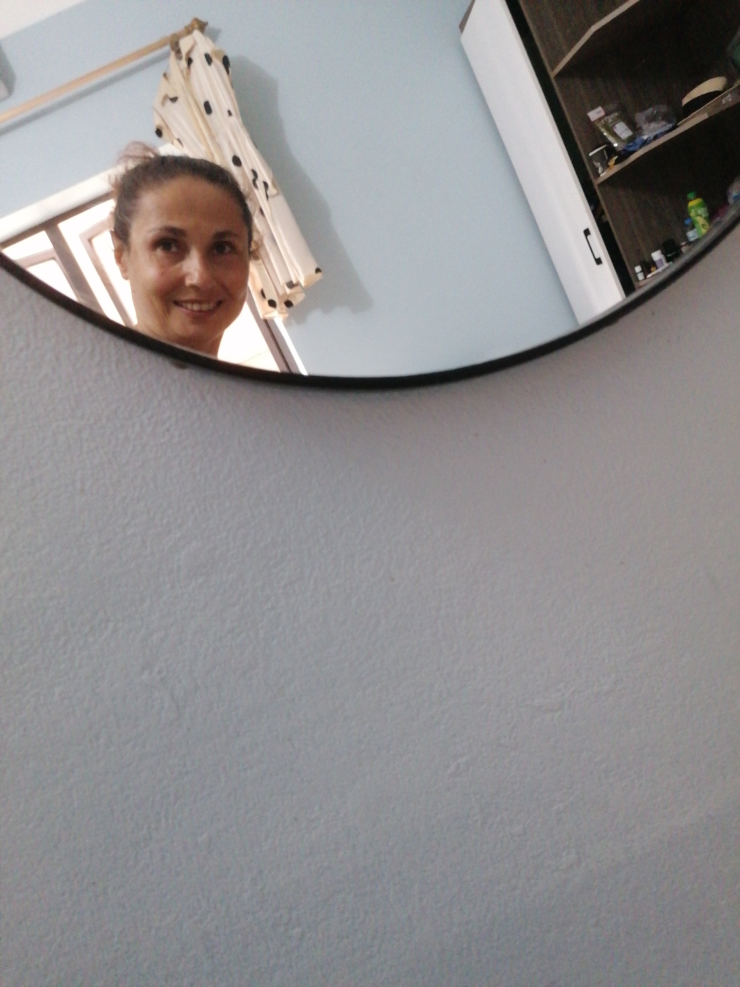 The image shows a partial view of a woman's face reflected in a round mirror. The woman is smiling. In the background, a window with light filtering through a white curtain with black polka dots is visible. The wall behind the mirror is light blue. A cabinet or shelf is visible in the upper right corner of the mirror's reflection, containing various items. The room appears to be in a residential setting. The location is Bai Chay Ward, Vietnam, based on the prompt.