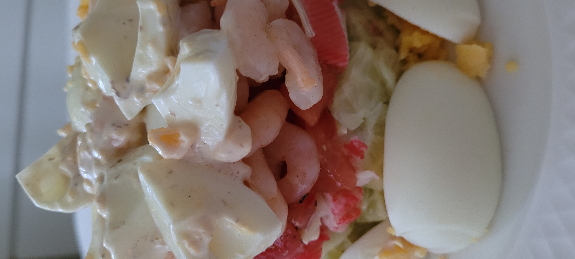 The image displays a close-up of a seafood salad, possibly a shrimp salad, served on a white plate. The salad appears to be layered, with chunks of hard-boiled egg, pink shrimp, red tomato pieces, and possibly some shredded lettuce or other greens visible. A creamy dressing coats the top layers of the salad. The focus is tight, capturing the textures and colors of the ingredients. There is no visible context of Riga, Latvia, or any other specific location.