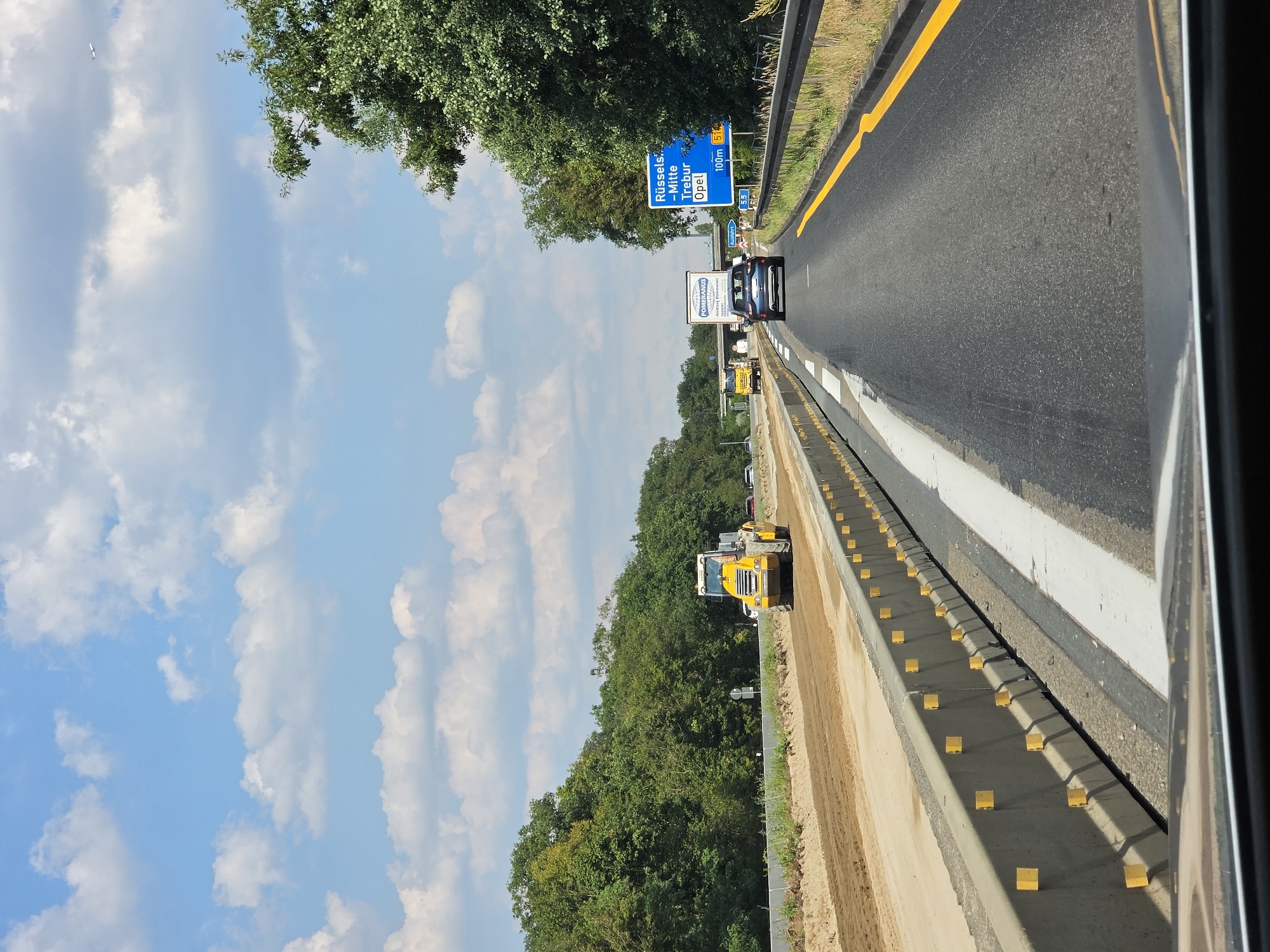 The image depicts a view from inside a vehicle traveling on a highway in Rüsselsheim am Main, Germany. On the left side of the highway, there is a construction zone with a yellow steamroller operating on a dirt surface. Concrete barriers with yellow reflectors delineate the construction area from the active lanes of traffic. On the right side of the highway, a blue road sign indicates "Rüsselsheim - Mitte, Trebur, Opel" and an exit at 100 meters. Another smaller blue sign displays "Ausfahrt 55". A dark blue car is traveling ahead in the right lane. The highway is bordered by trees and vegetation on both sides. The sky is blue with scattered white clouds.