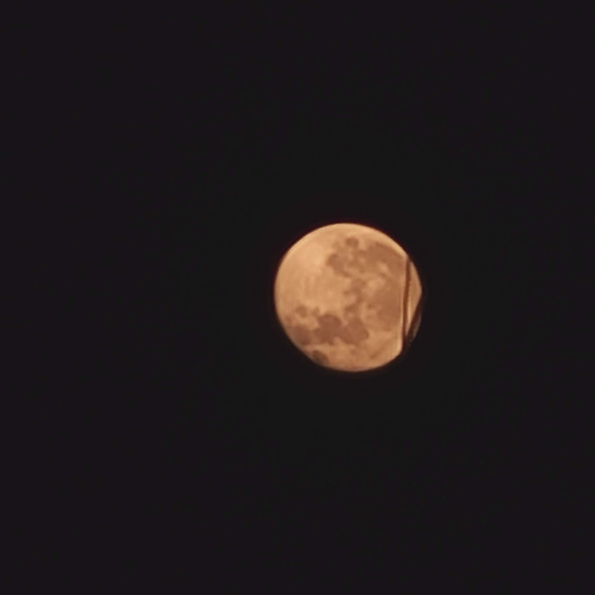 The image shows a full moon against a dark sky. Part of a structure (possibly a wire or pole) is visible in front of the moon on its right side. The image was taken at night in an unidentified location in San Fernando del Valle de Catamarca, Argentina. There are no people or other subjects present in the frame.