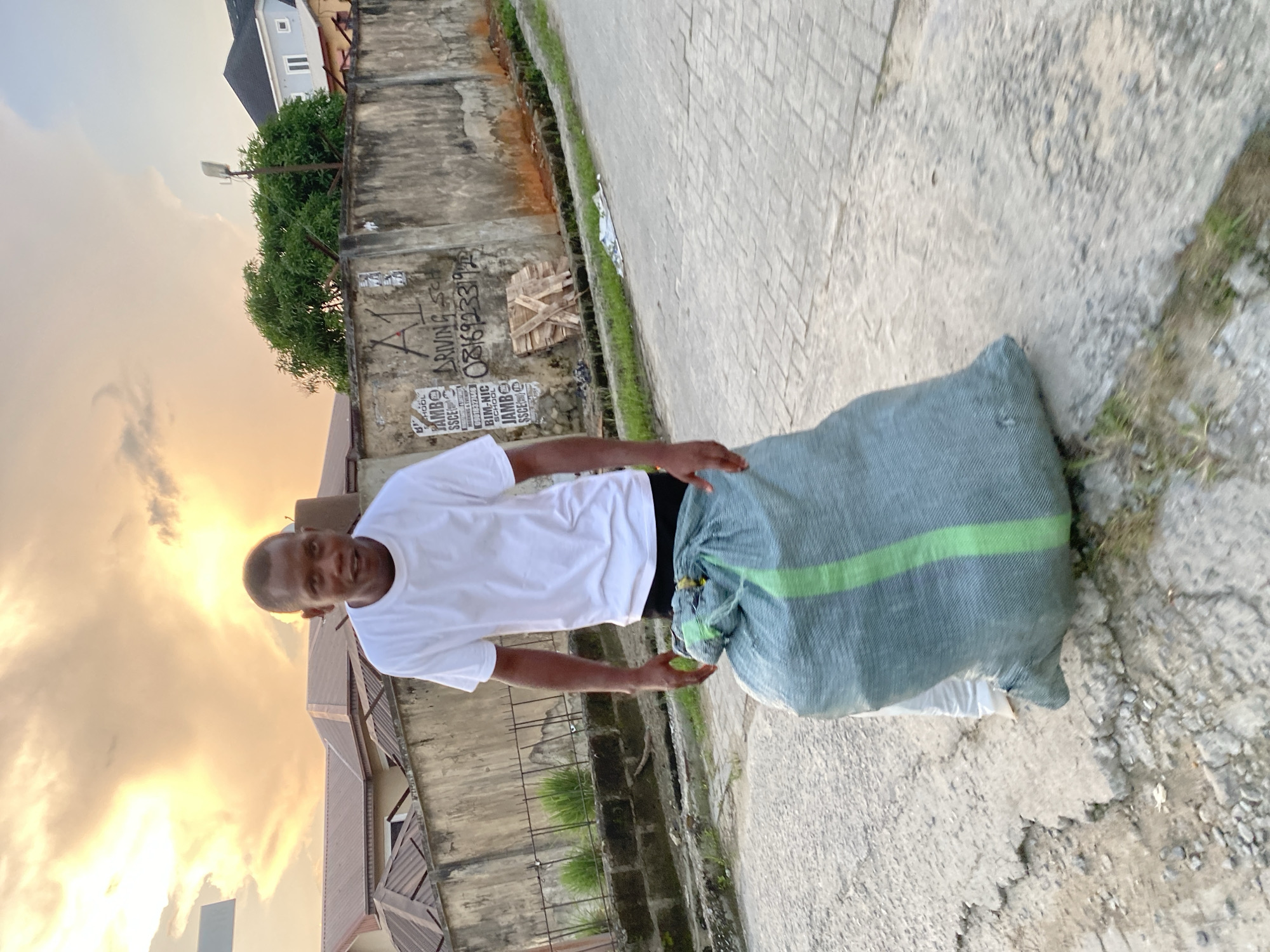 A man, appearing to be of African descent, stands on a paved road with a large, light-blue duffel bag with a green stripe in front of him. He is wearing a white t-shirt and black shorts. The bag is cinched at the top with a yellow tie. The man is looking towards the camera with a neutral expression. Behind him, a concrete wall is covered in graffiti, including the words "A1 DRIVING SCHOOL" and a phone number. To the left of the wall are houses with brown roofs and a lush green tree. The sky above is filled with dramatic, golden clouds, suggesting either sunrise or sunset. The scene appears to be in a residential area of Ado, Nigeria.