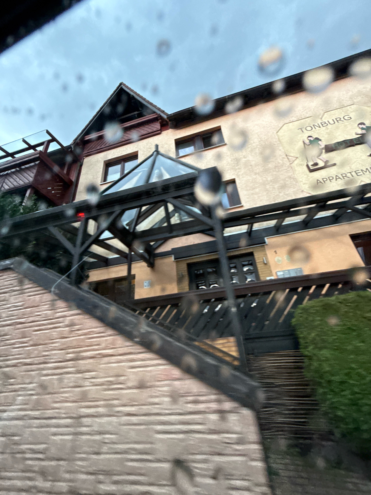 The image shows the facade of the "Tonburg Hotel & Appartements" located in Bad Sachsa, Germany. The building has a light-colored exterior with a prominent sign featuring the hotel's name and a graphic representation of two figures. The architecture includes multiple windows and a modern glass-covered walkway or entrance. A steep, brick-covered slope leads up to the building. The foreground of the image is blurred and shows water droplets, suggesting the photograph was taken through a wet window, possibly from a moving vehicle. The sky above is overcast.
