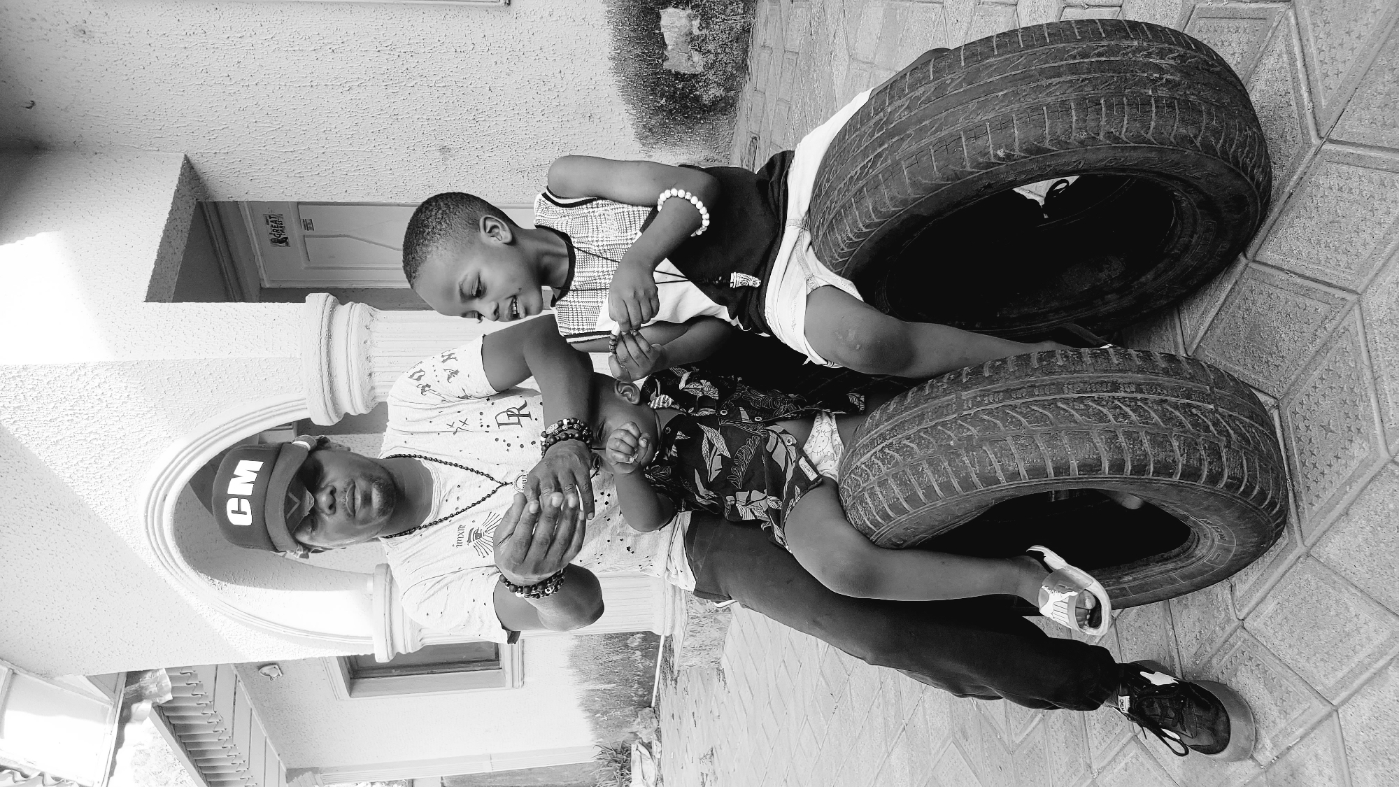 This black and white image captures an outdoor scene in Sabon Tasha, Nigeria, depicting an adult male interacting with two young children during the daytime. The setting appears to be a paved patio or courtyard, featuring distinctive diamond-patterned tiling on the ground.
In the foreground, an adult male is seated or leaning, wearing a dark cap with the letters "CM" clearly visible on the front, a light-colored patterned t-shirt, a long beaded necklace, and multiple beaded bracelets on both wrists. He looks down with a focused expression, cradling an infant in his left arm and holding the infant's hands with his right.
To the adult's right, a young boy leans in, engaging with the infant. He wears a patterned top, light-colored shorts, and a white beaded bracelet on his right wrist. The boy has a bright smile on his face as he looks at the infant, extending his hands towards the baby's hands in a playful gesture. The infant, wearing a patterned romper, is partially visible, lying on their back, held securely by the adult.
Two dark, worn vehicle tires are prominent objects on the paved ground to the right of the subjects. The background shows architectural elements, including a white fluted pillar, a textured wall, and a door with a small sign above it that reads "GREAT" and possibly another word below it. The bright lighting indicates it is daytime, and the overall mood of the image is one of warm family interaction and tenderness.