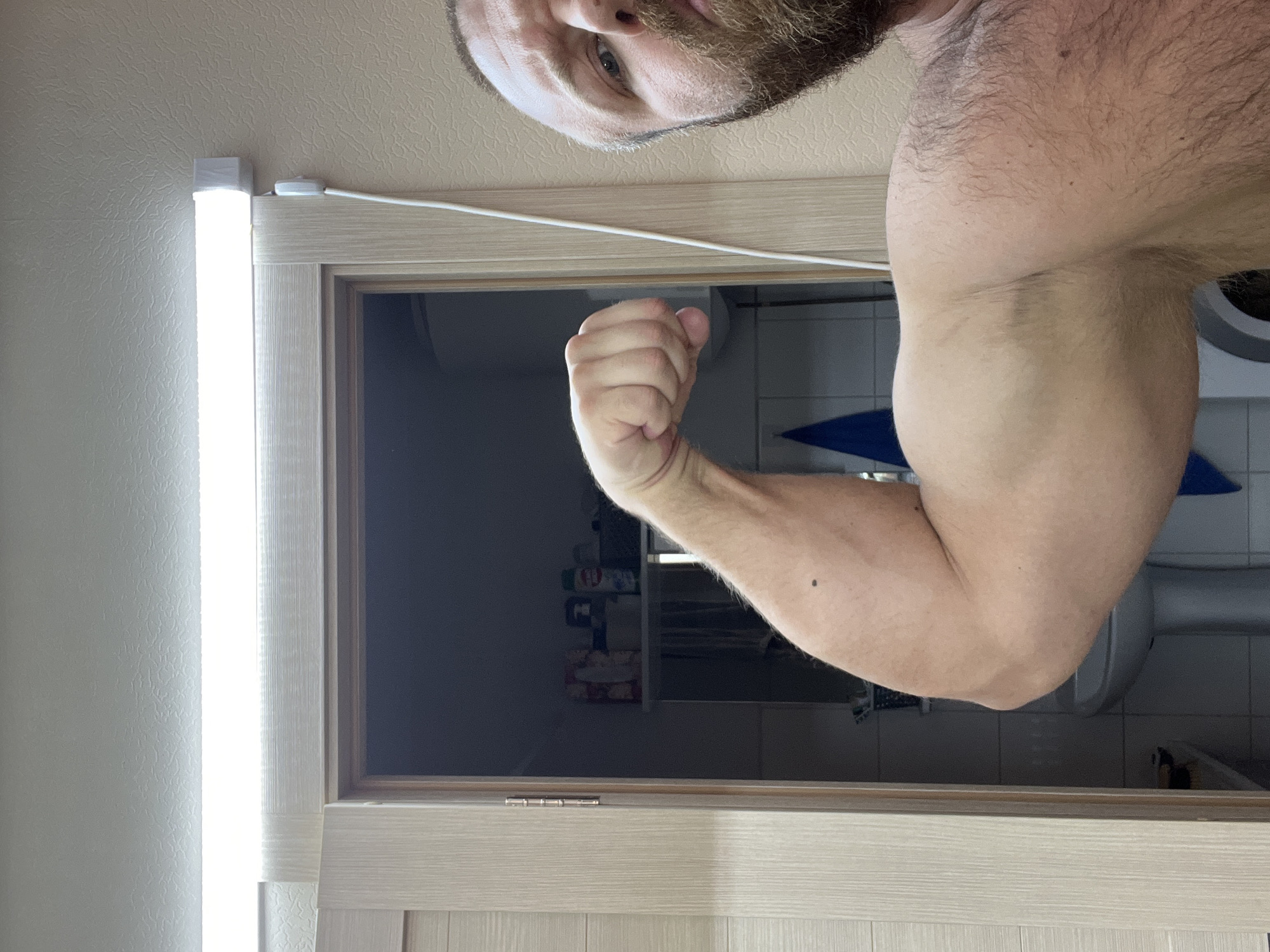 A man is taking a selfie in a bathroom. He is holding his arm up and flexing his bicep. The background shows a toilet, sink, and some toiletries. A bright light fixture is adjacent to a door frame. The location is Noviy, Russia.
