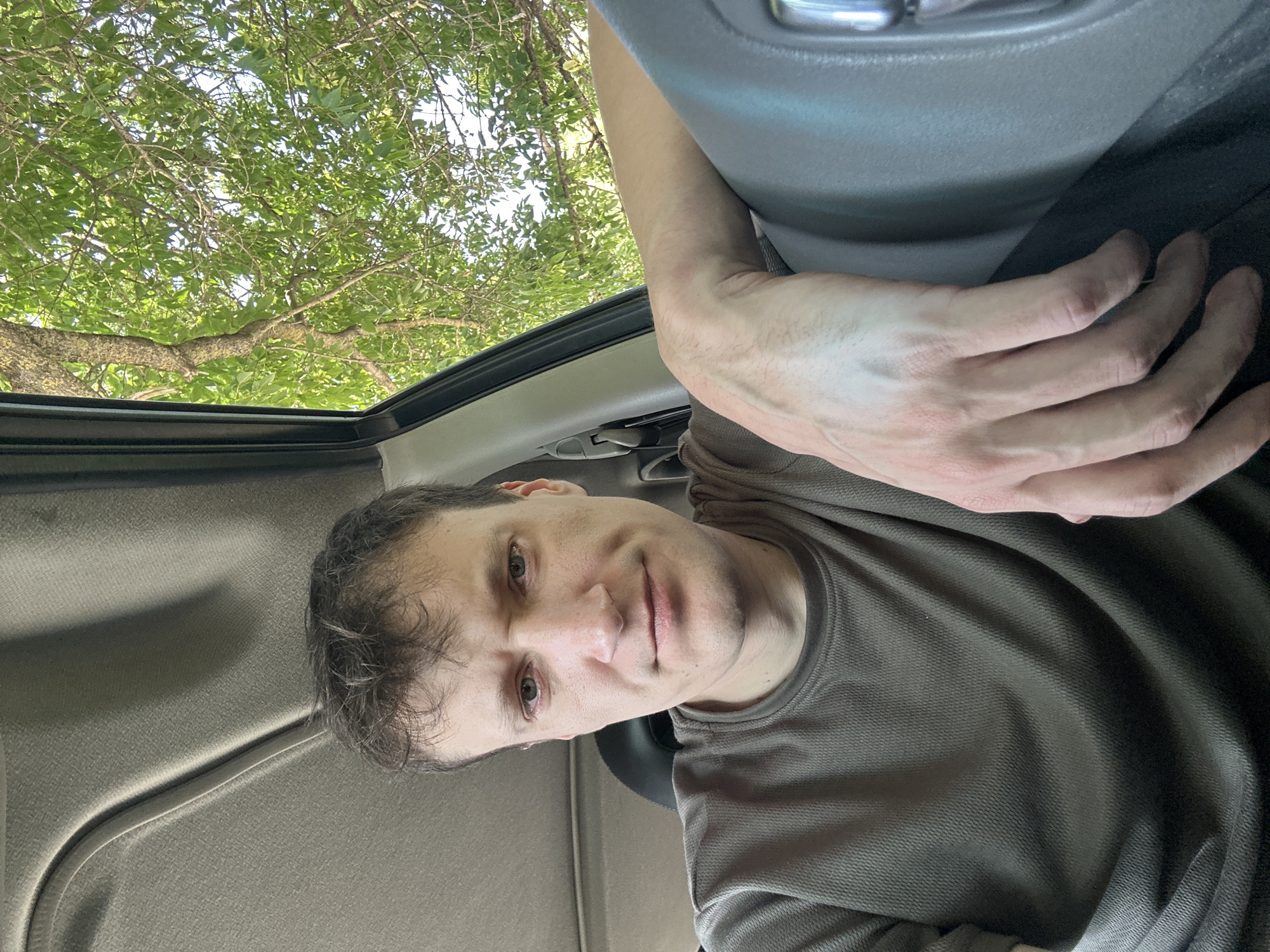 A Caucasian male with short, dark, messy hair is sitting in the driver's seat of a car. He is wearing a short-sleeved, taupe t-shirt. His right arm is resting on the center console of the car. The interior of the car is visible, including the headliner and a portion of the steering wheel. Outside the car, through the open window, green foliage and tree branches are visible against a bright sky. The man's facial expression is neutral, with his eyes looking directly at the camera. The image appears to be a selfie taken from the driver's perspective. The location is the city of Makiv, Ukraine.