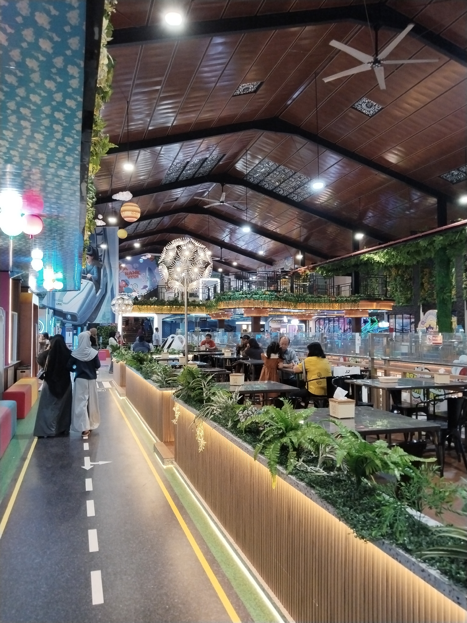 This image depicts the interior of a brightly lit, multi-level indoor entertainment venue or dining establishment in Kecamatan Pati, Indonesia. The scene is bustling with activity, suggesting it's a popular spot.
In the foreground, two individuals wearing hijabs walk along a pathway marked with dashed white lines and a yellow border, reminiscent of a road. The pathway is flanked by planters filled with lush green foliage and illuminated by warm LED strip lighting embedded in a slatted wooden wall.
The venue features a high, vaulted ceiling with exposed wooden beams and panels, fitted with multiple industrial-style ceiling fans and recessed lighting. Decorative elements are suspended from the ceiling, including spherical pendant lights, globe-like lanterns resembling planets, and a large, dandelion-shaped light fixture.
The midground reveals seating areas with tables and chairs, where other patrons are seated, some engaged in conversation or dining. A second level of the venue is visible above, with more seating and a railing, also adorned with hanging greenery. In the background, there are various entertainment attractions, including what appears to be a VR experience or simulator with a large mural depicting a person in a VR headset. Neon signage and colorful lights suggest further attractions or shops.
The overall ambiance is lively and inviting, with a blend of natural elements like plants and modern architectural features. The lighting indicates it is likely evening or indoors with artificial lighting dominating the scene. No specific weather conditions are discernible, but the indoor setting makes it irrelevant. The decor and signage (though mostly illegible) hint at a family-friendly entertainment complex.