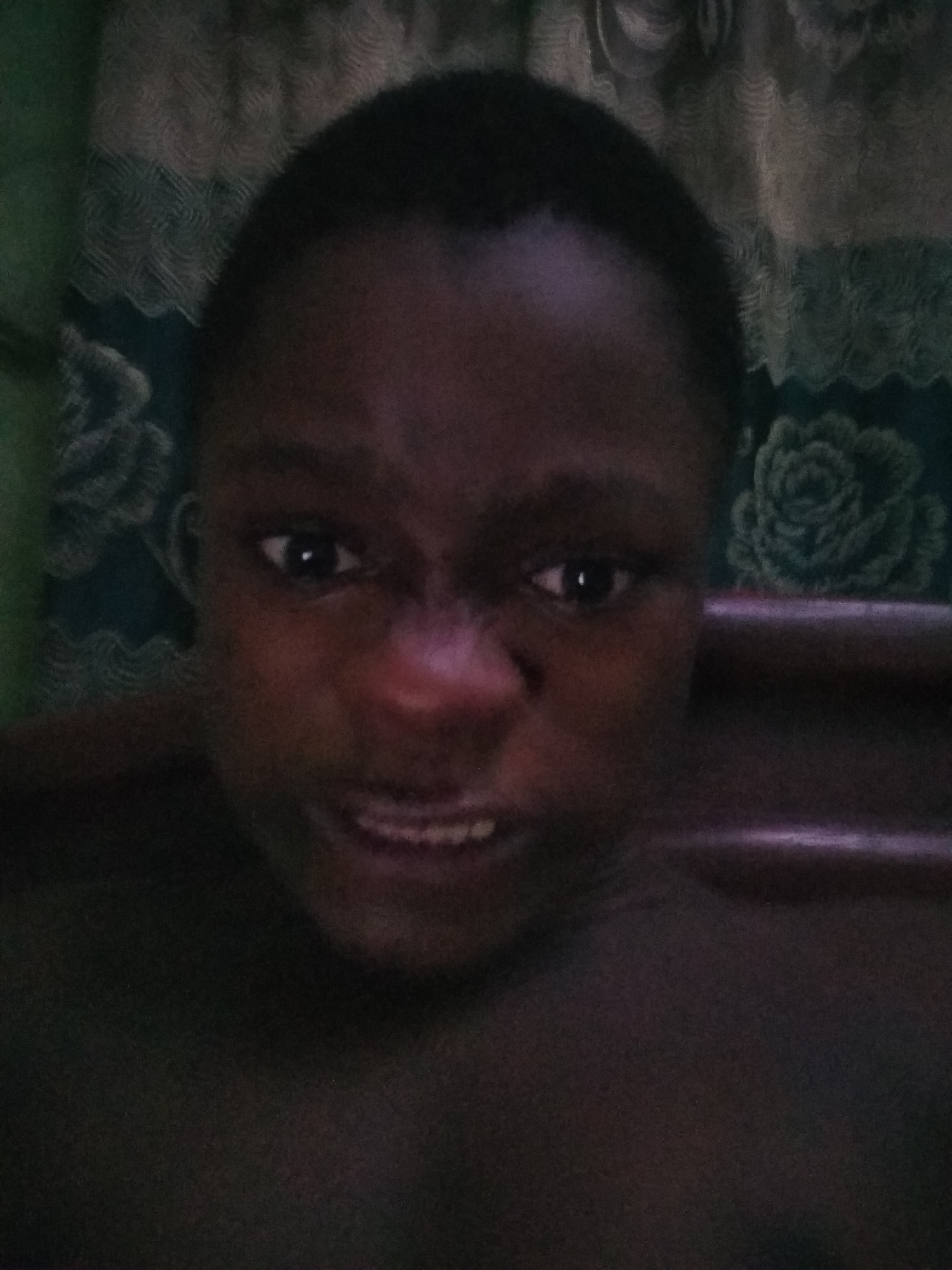 A close-up photograph depicts a dark-skinned person with a close-cropped hairstyle. The background shows a patterned curtain and a wooden object, potentially a bed frame. The image appears to be taken in a low-light environment. The facial expression is tense, with slightly bared teeth. The location is assumed to be Shangisha, Nigeria, but there is no confirming visual information in the image.