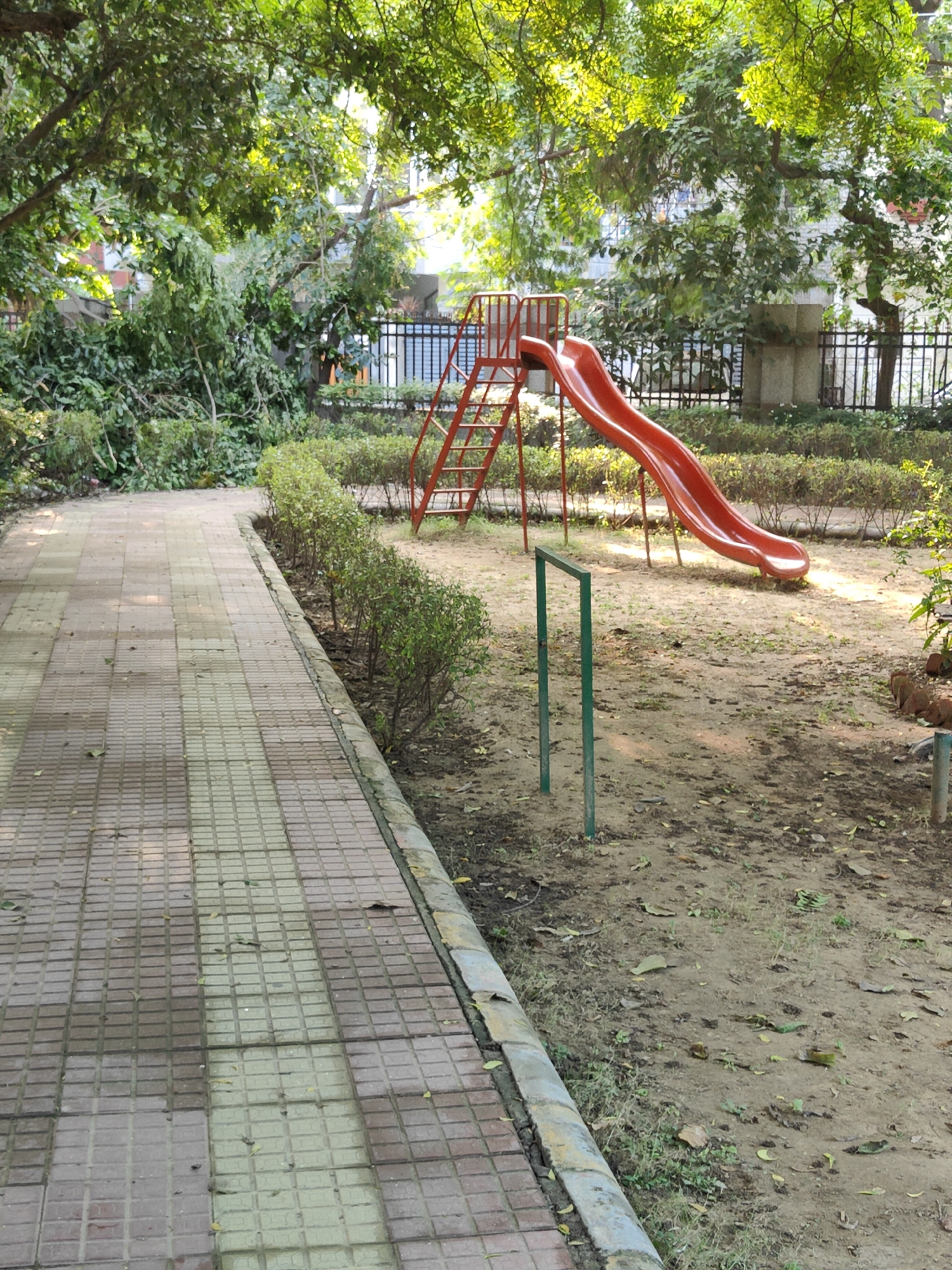 A red metal slide with a yellow ladder is positioned in a park on a sandy surface. The slide is angled to the right, with the top of the ladder on the left. A low, manicured hedge runs parallel to a tiled walkway, which is in the foreground and occupies the left side of the frame. In the background, a black metal fence encloses the park, with trees and foliage visible beyond. The scene is bathed in natural light, suggesting it is daytime. This park is located in Gurgaon, India.