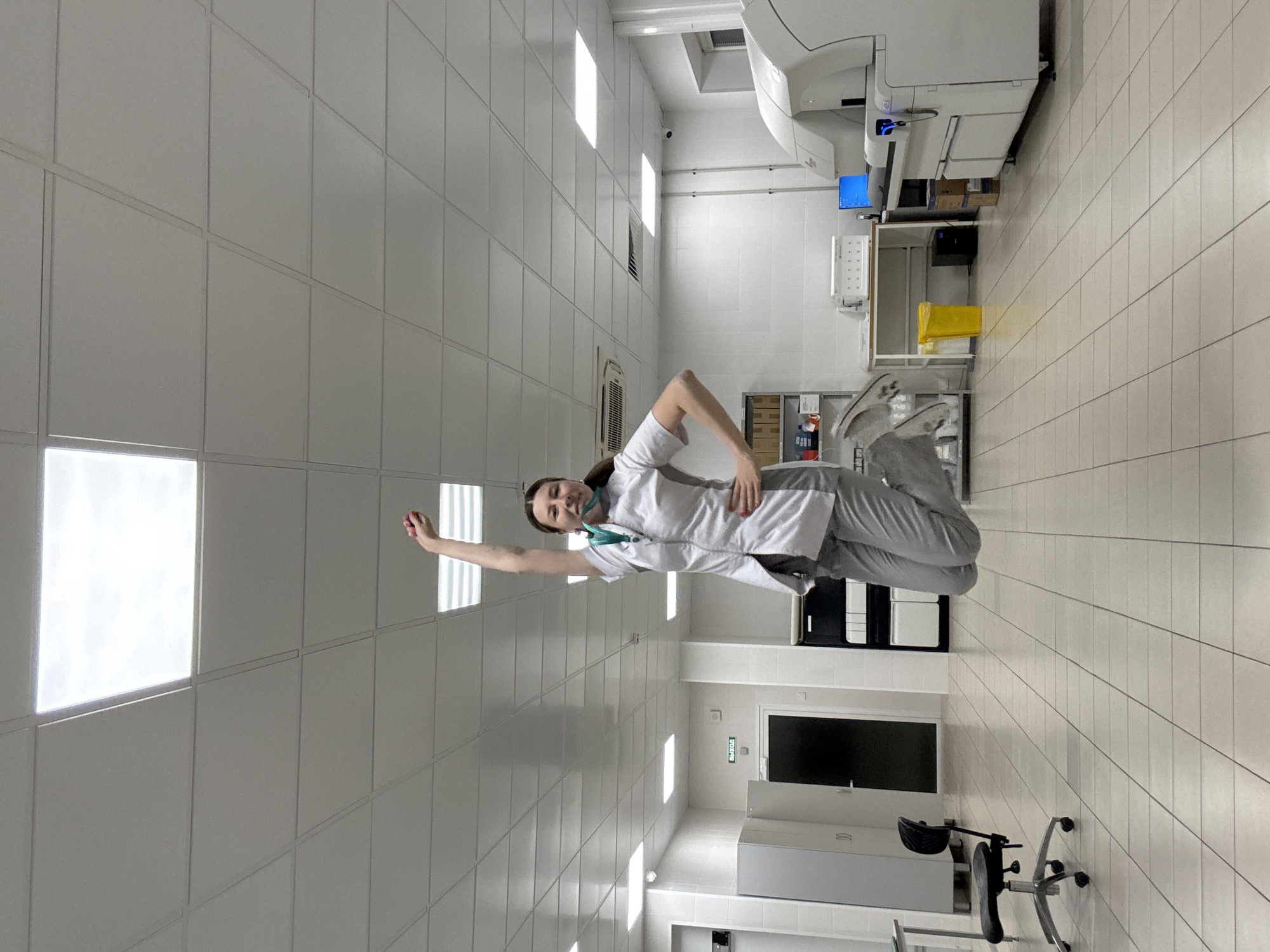 A woman in a white lab coat and grey pants is jumping in the air in a well-lit room. She is wearing a green face mask and has one arm raised in the air, holding a small red object. The room appears to be a laboratory or medical facility, with various pieces of equipment visible, including a large white machine on the right side of the image, a desk with a computer monitor, and a yellow trash can. The floor is tiled, and the ceiling has fluorescent lighting panels. There is a door with a green "EXIT" sign on the left wall. The image does not contain any information to determine its location in Moscow, Russia.