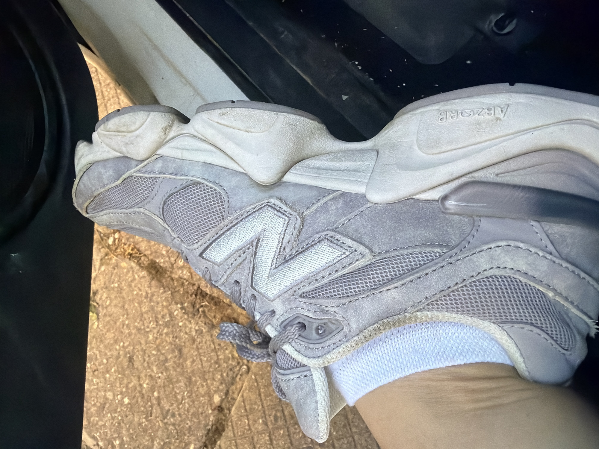 A close-up shot depicts a grey New Balance sneaker being worn, with a white sock visible. The sneaker is positioned near the open door of a vehicle, possibly a car. The background shows a glimpse of pavement outside. The location is Unknown, Unknown.
