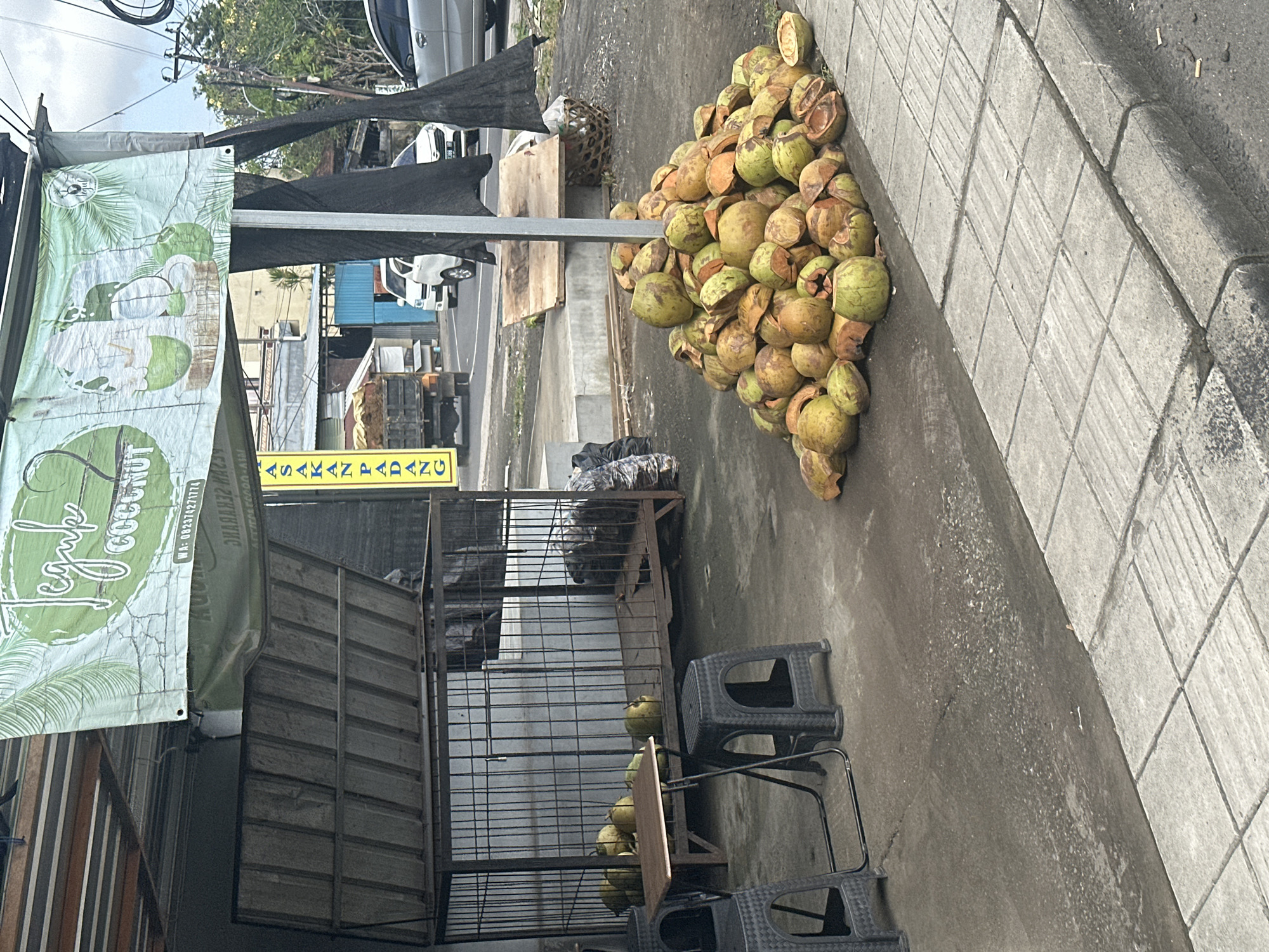 A roadside coconut stand in North Kuta, Indonesia, is depicted. The stand features a large banner with the text "Teguk Coconut" and images of coconuts. In front of the stand, a pile of cut coconuts is visible, with more coconuts inside a metal cage. A small wooden table and two plastic chairs are also present. In the background, vehicles are parked and driving on the street, and buildings line the road. The scene is set on a paved area with a sidewalk.