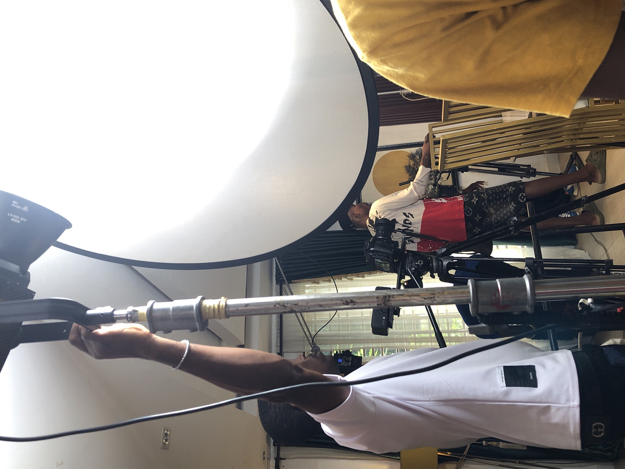 An indoor scene depicts a film or photoshoot in progress, featuring two men and various production equipment. The image is oriented vertically, showing the subjects and equipment from a side perspective.
In the foreground, a man with a dark complexion, wearing a white short-sleeved t-shirt and dark shorts, extends his arms upwards, holding a long, silver-colored boom pole, likely for audio recording or lighting. A white beaded bracelet is visible on his right wrist. His white t-shirt has a small black rectangular label on the chest.
Further back and to the right, a second man, also with a dark complexion, operates a professional video camera mounted on a tripod. He wears a long-sleeved white shirt with red and black graphic designs and patterned shorts featuring the distinct "LV" monogram, indicative of Louis Vuitton. Text on his shirt includes "SON OF GOD" in red and "MINDS" in black, with "ANTI HERO" and "SIMONS" also partially visible. He is wearing light green Crocs-style shoes.
Dominating the upper left portion of the frame is a large, brightly illuminated white circular softbox, providing key lighting for the setup. In the upper right background, a gold-colored slatted bench or prop is visible, along with a draped yellow piece of fabric. Black cables traverse the scene, connecting to equipment. A window with blinds can be seen in the background, suggesting natural light is also present, though the primary lighting seems to be artificial. The overall setting appears to be a studio or a room adapted for professional media production. The men are focused on their tasks, indicating a concentrated working environment.
