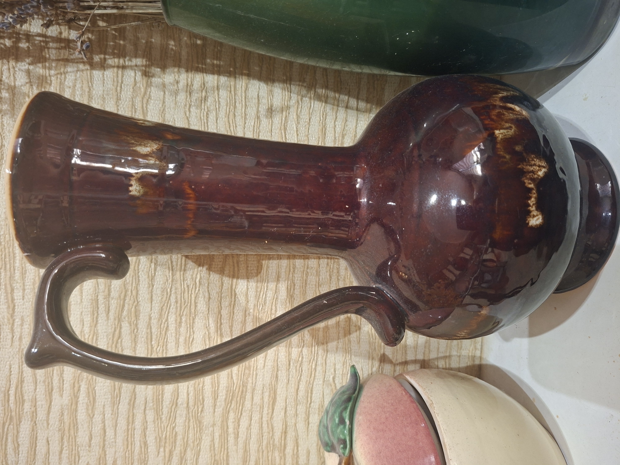 A dark brown ceramic pitcher with a glossy finish stands on a white surface. The pitcher has a large, curved handle and a wide opening. To the left of the pitcher is a portion of a light-colored ceramic bowl topped with a pink object and a green leaf-like decoration. To the right of the pitcher is a tall, dark green glass vase containing dried flowers. The background is a textured, light-colored wall with horizontal lines. The scene appears to be indoors, possibly in a home or shop, with natural light casting shadows from the objects.