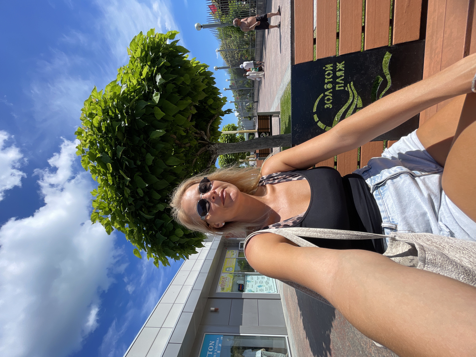 A blonde woman with sunglasses and a leopard-print tank top sits on a wooden bench in the city of Anapa, Russia. She is holding a light-colored tote bag over her shoulder. In front of her is a black sign with green lettering that reads "ЗОЛОТОЙ ПЛЯЖ" (Golden Beach). Behind her, a path lined with trees and benches leads toward a fence. Several people are visible walking in the distance. The sky is blue with scattered clouds, and sunlight casts shadows on the ground.