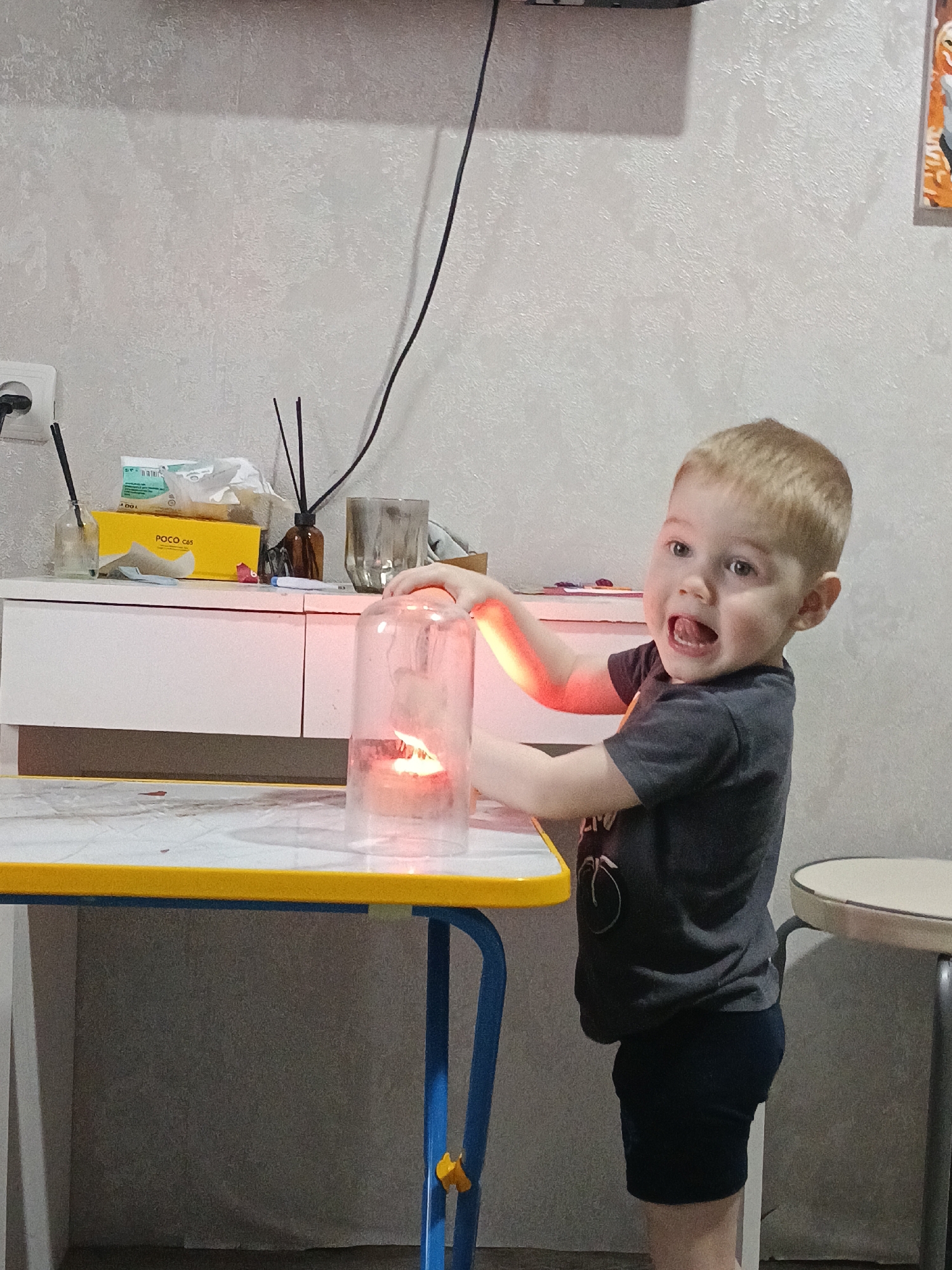 A young child stands next to a table in an interior location, possibly a home. The child has short blonde hair and is wearing a dark gray t-shirt with a design on it, and dark blue shorts. The child has their mouth open and appears to be speaking or making a noise. On the table, there is a lit candle inside a glass dome. The table has a white surface with a yellow edge, and blue legs. Behind the table is a white cabinet with various items on top, including boxes, glassware, and incense sticks in a brown bottle. The wall behind the cabinet is light gray. In the background, near the top of the image, there is a black electrical cord hanging down. To the right of the child, there is a small white stool with a metal frame. The child seems to be interacting with the candle and glass dome on the table. The city of Chaikovsky, Russia, is mentioned as the location.