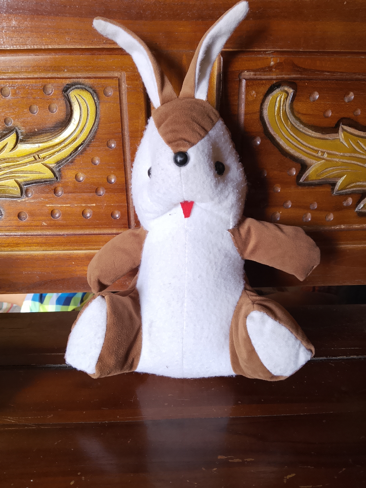 A stuffed rabbit toy with white and brown fur sits on a wooden surface. The rabbit has long, floppy ears, black eyes, and a small red mouth. It is positioned in the center of the frame, facing forward. The wooden surface appears to be part of a bench or table with decorative carvings. The background is also wooden and features ornate, golden designs. The scene is indoors.