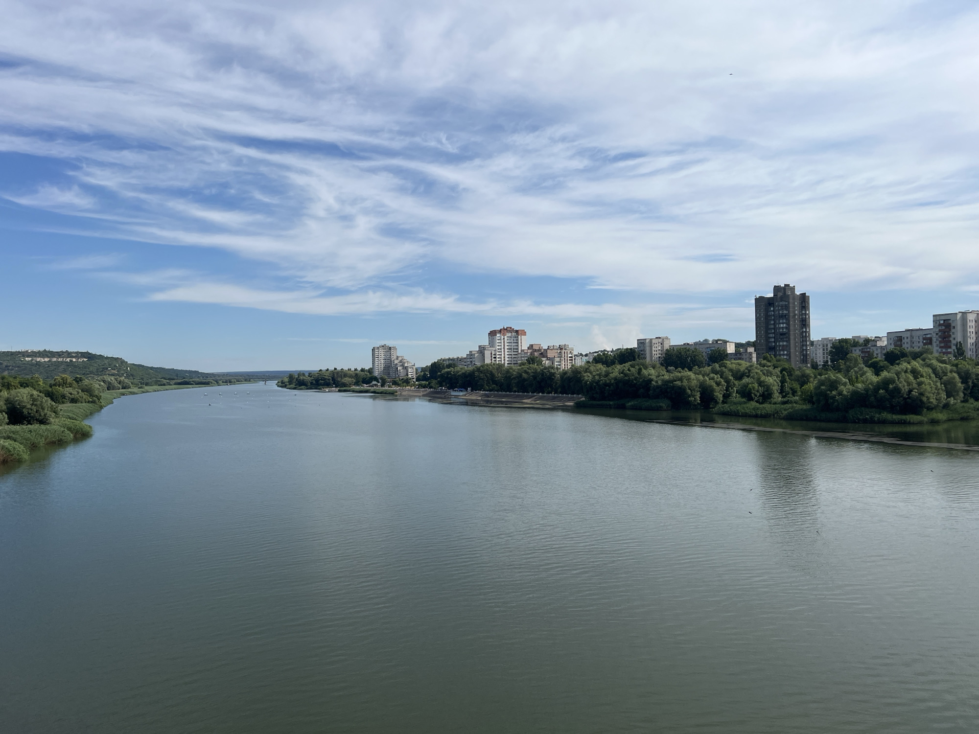 A wide, calm river flows through a city, with buildings and trees lining the banks under a partly cloudy sky. The cityscape features several apartment blocks of varying heights and architectural styles, interspersed with lush green trees. The water's surface is disturbed by gentle ripples, reflecting the sky and surrounding scenery. In the distance, a bridge spans the river. The scene appears to be set in the city of Rezina, Moldova. There are no visible people or specific actions taking place. The environment is predominantly natural with urban development along the riverfront.