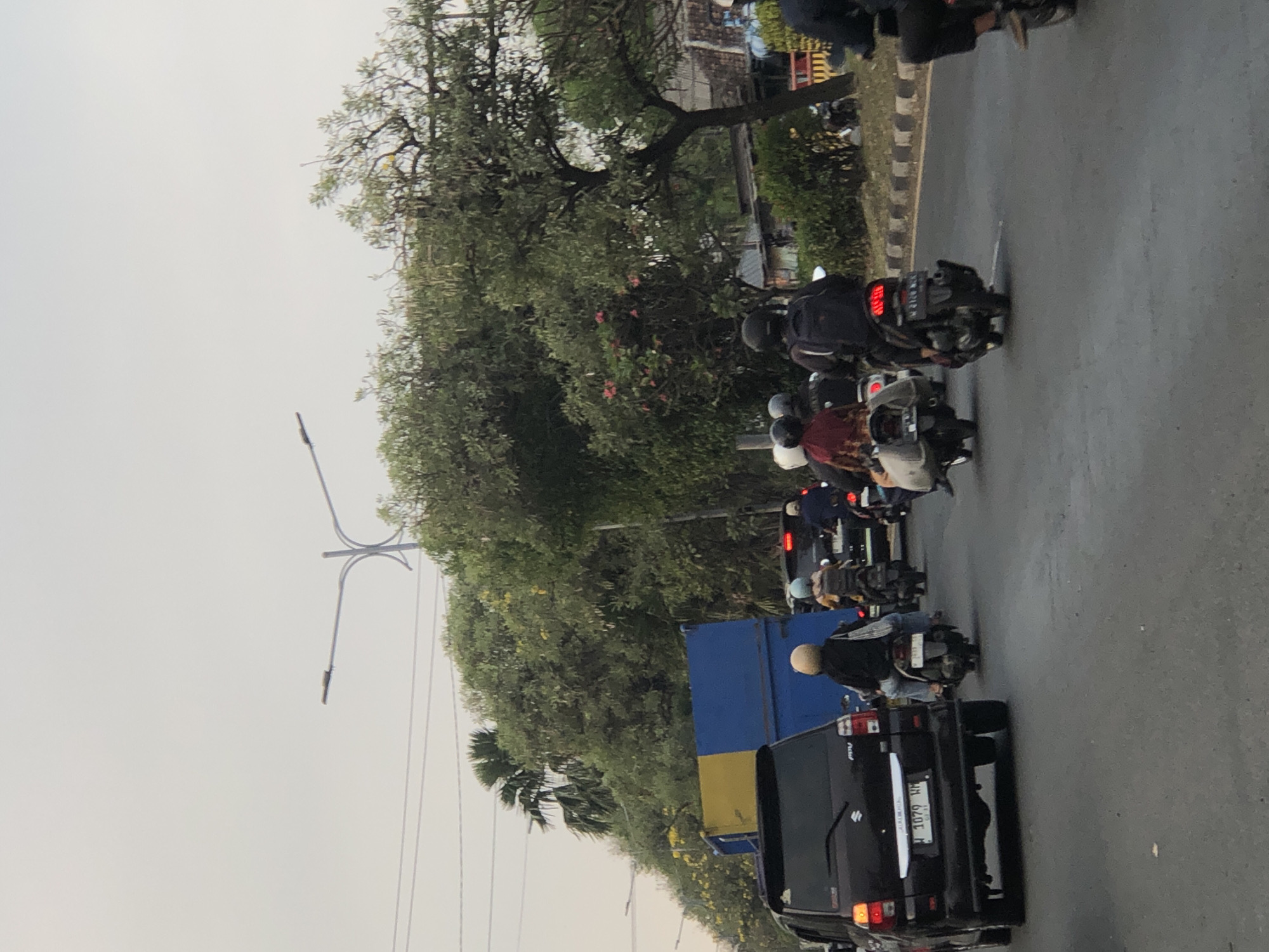 The image depicts a traffic scene on a street in Ngaban, Indonesia, likely during daylight hours with overcast skies. In the foreground, a black SUV is visible on the left, with a license plate reading "B 1079 WH". Multiple individuals are riding motorcycles, primarily in the middle and right sections of the frame. Some are wearing helmets, while others are not. They are navigating through traffic. Behind the SUV, a large blue and yellow truck is present. The background features lush green trees and foliage lining the road, with a street lamp visible in the upper left quadrant. The road surface appears to be asphalt.