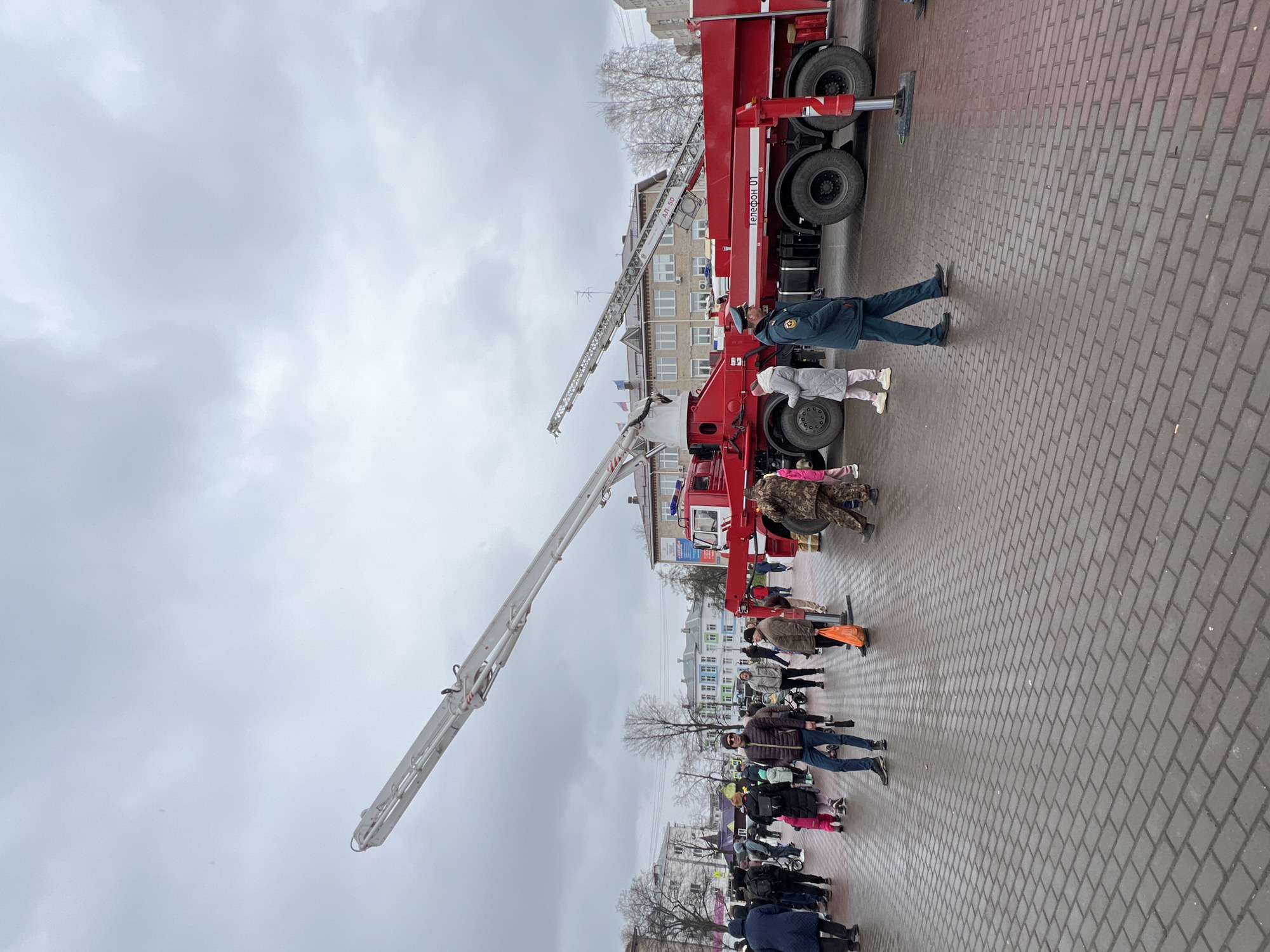 The image depicts a red fire engine with an extended aerial ladder parked on a paved brick plaza, likely in the city of Chaikovsky, Russia. People of various ages are visible in the surrounding area, suggesting a public gathering or demonstration. One person in a dark uniform near the fire truck seems to be speaking to two children. The overcast sky suggests a cool day. Buildings are visible in the background.