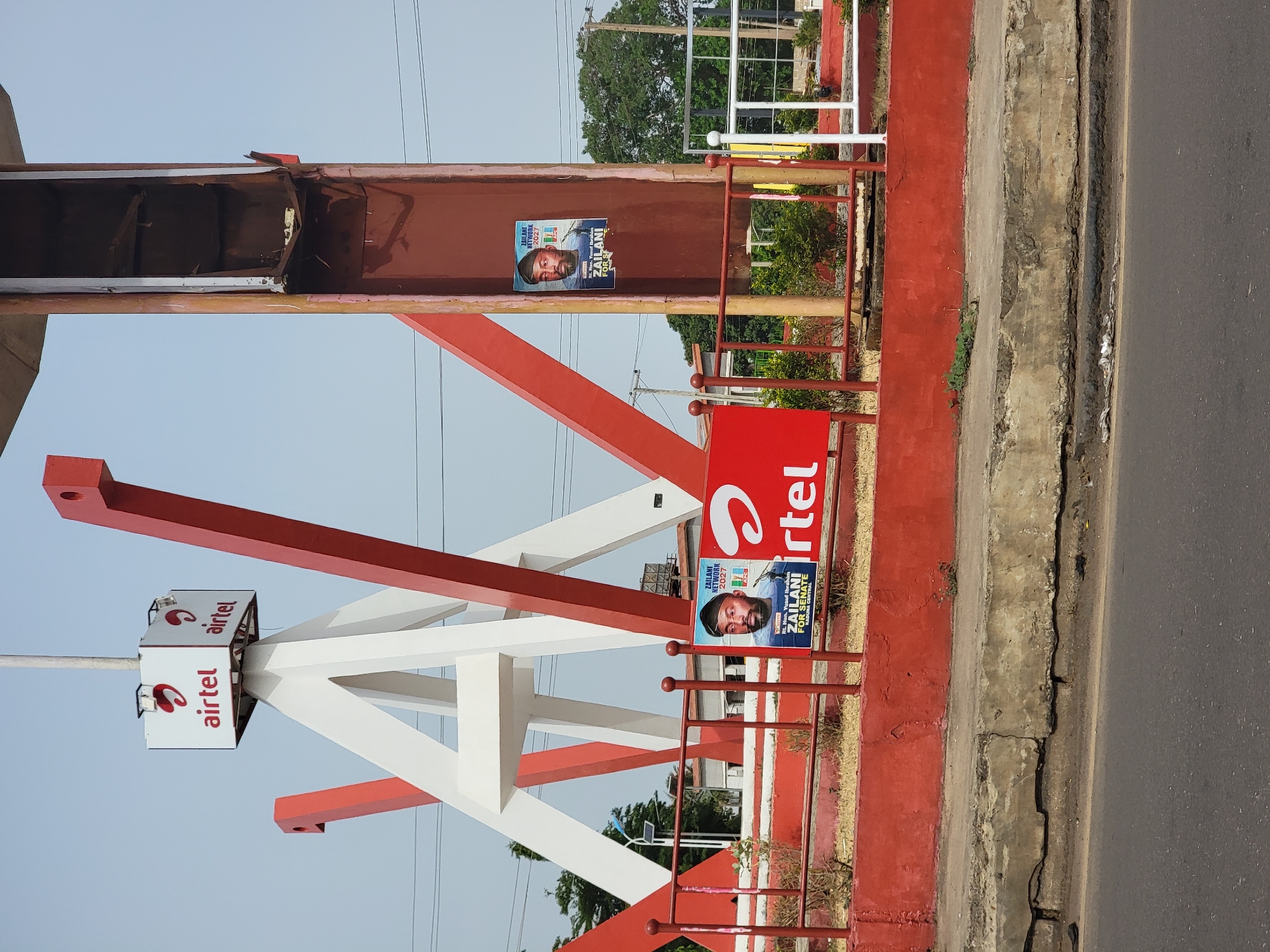 The image depicts a roadside scene in Barnawa, Nigeria, featuring an Airtel telecommunications sign with accompanying support structure painted in red and white. Political posters for "Zailani for Senate" are affixed to the structure, promoting a candidate for the 2027 elections. The surrounding environment includes a road with concrete edging and vegetation. The metal beams of the sign structure show signs of wear and deterioration. A fence with red posts and white rails is also visible.
