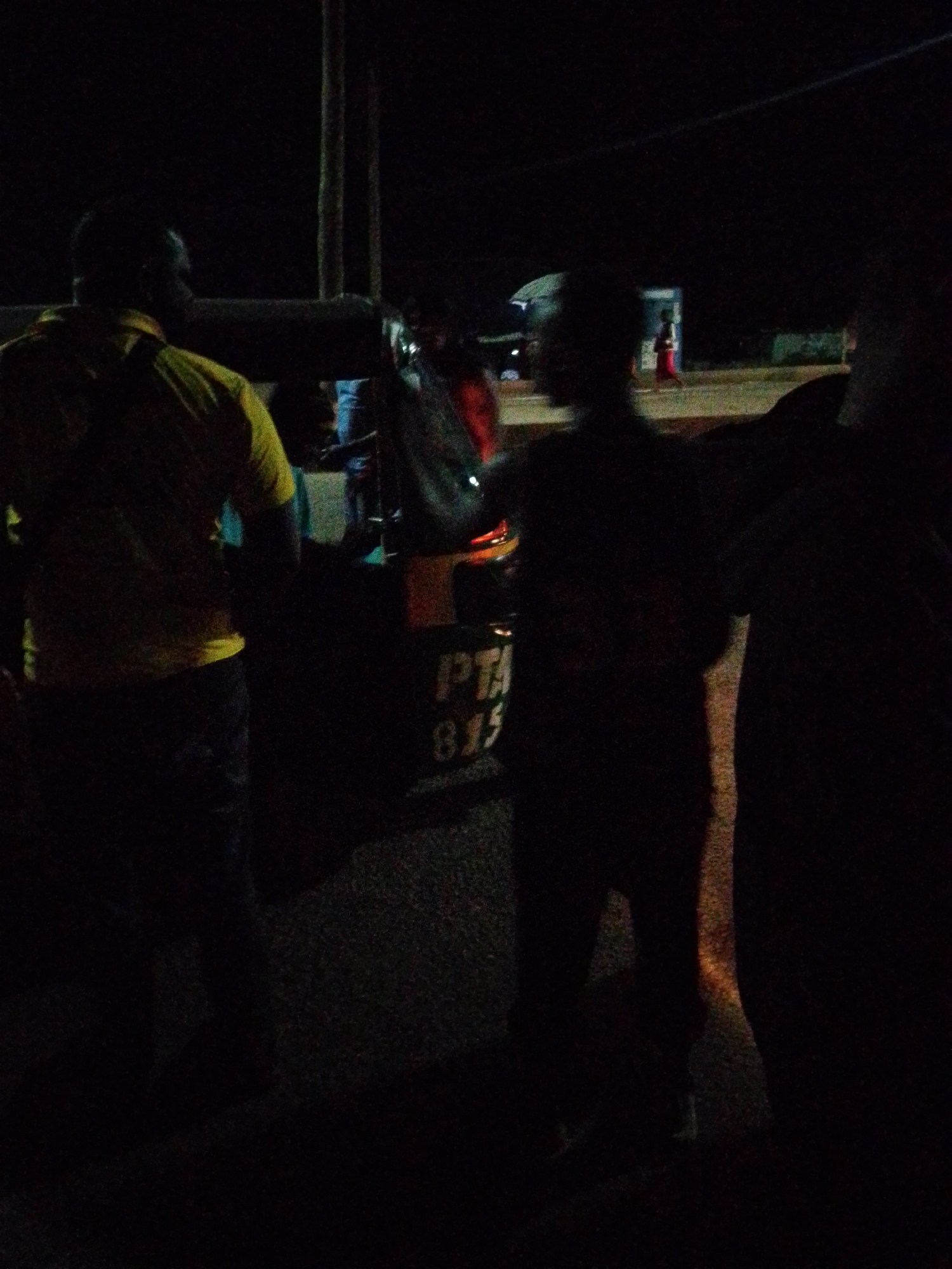 In the city of Bukuru, Nigeria, at night, a person stands in the foreground with their back to the camera, wearing a yellow shirt and dark pants. To their right, another individual is silhouetted against the darkness. Behind them, a vehicle or cart is visible, with the letters "PTA" and the number "815" painted on its side. The background is dark, suggesting a rural or less developed area with some faint lights and structures in the distance.