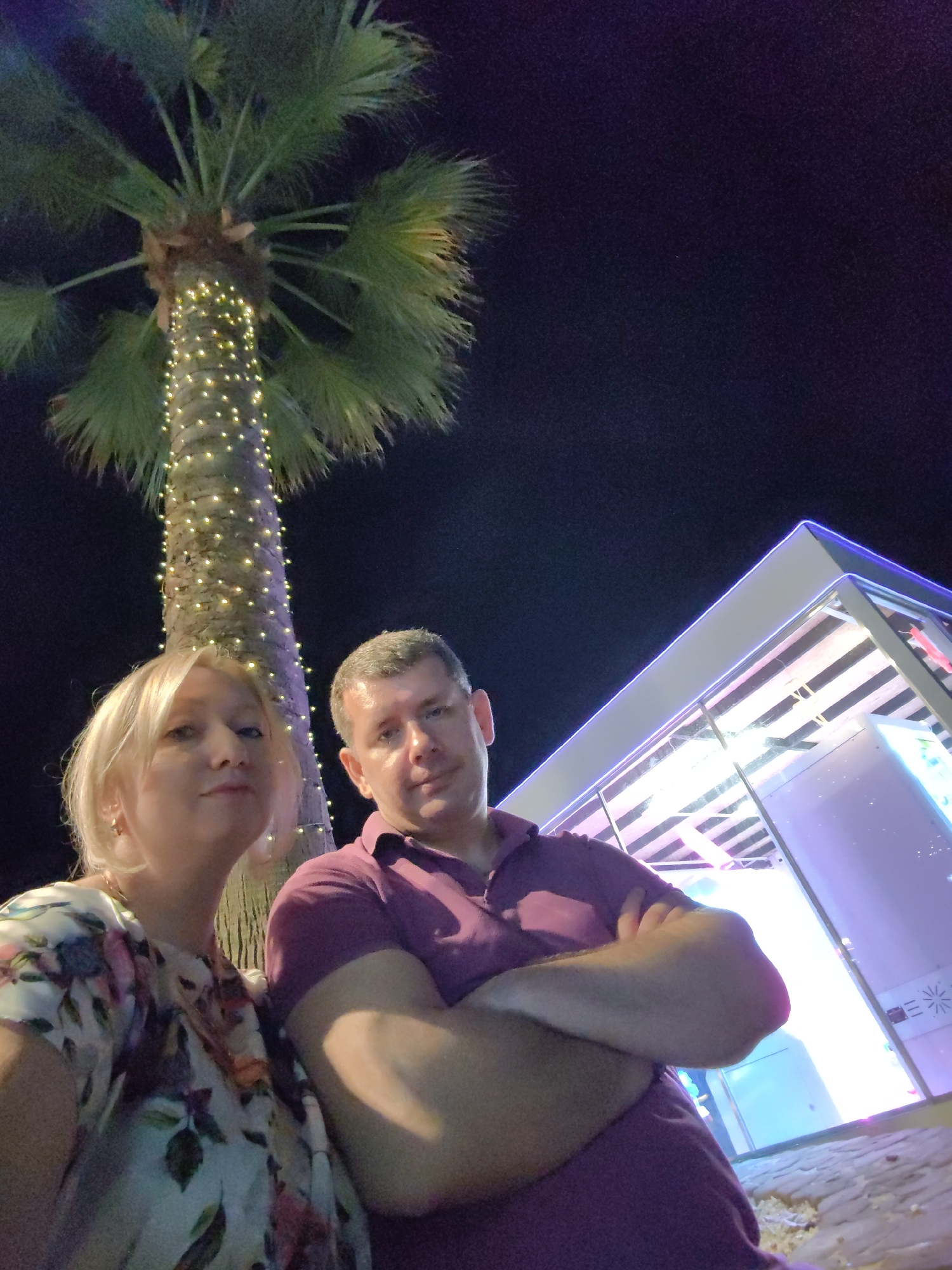 Two people, a woman and a man, are posing for a photo at night in Sochi, Russia. The woman is on the left, with blonde hair and a floral top. The man is on the right, wearing a purple polo shirt with his arms crossed. Behind them, a palm tree is decorated with string lights, and a modern building with neon lighting is visible. The sky is dark, indicating it is nighttime. The ground appears to be paved with cobblestones.