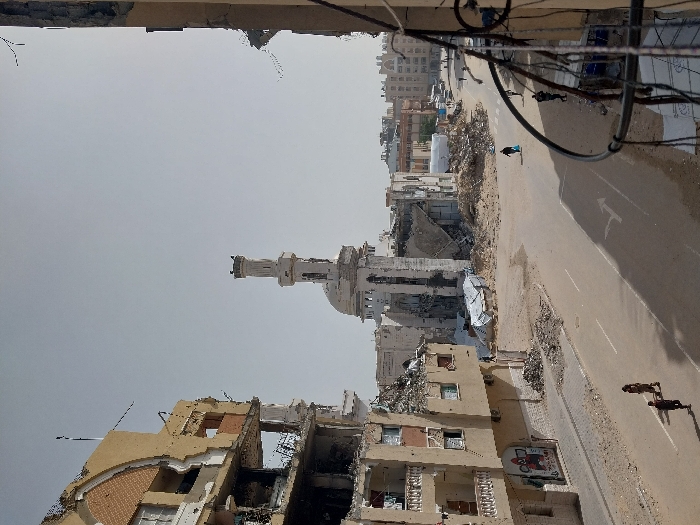 The image depicts a scene of extensive destruction in an urban area of Khan Yunis, Palestinian Territory, under a hazy sky. In the foreground-left, a multi-story building, likely residential or commercial, shows severe structural damage; its exterior walls are partially collapsed, exposing multiple floors with visible debris and mangled structural elements. Directly across the street, in the mid-ground, stands a heavily damaged mosque with its minaret largely intact but the dome and surrounding structures significantly compromised and partially collapsed, surrounded by large piles of rubble.
A paved road runs through the center of the frame, showing a faded white arrow painted on its surface. The road itself is partially obscured by sand and scattered building debris. Three distinct individuals are visible on the street: two are walking together towards the lower-left corner of the frame, one in dark attire and the other in a patterned dress. A third individual, wearing dark clothing and carrying a blue object, walks across the street from right to left in the mid-ground. Additional indistinct figures are visible further down the street.
The general environment indicates widespread devastation, with numerous piles of concrete, rebar, and other building materials scattered across open areas and along the roadside. Other partially destroyed buildings and outlines of structures are visible in the background, extending into the distance. A black metal railing is present in the lower right corner, suggesting the photograph was taken from an elevated vantage point such as a balcony or window.