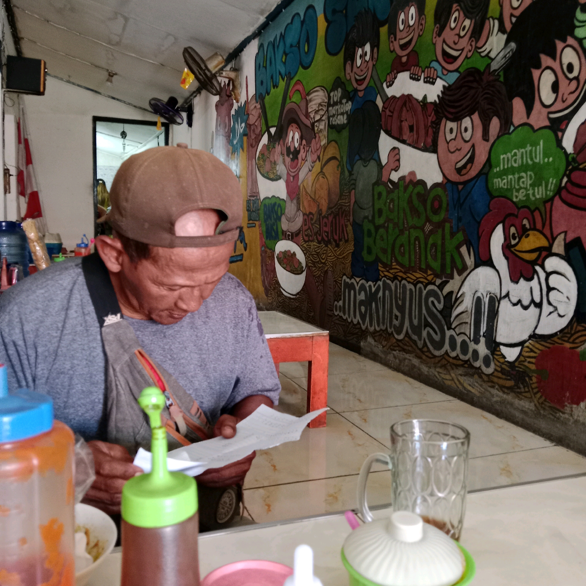 The image depicts a man wearing a brown baseball cap and a grey t-shirt, sitting at a table in what appears to be an outdoor food stall or small restaurant in Mandiraja, Indonesia. He is looking down at a piece of paper he is holding, possibly an order or bill. In front of him, on the table, are various condiments in plastic bottles, a glass mug, and a white container with a lid. The background features a colorful mural on the wall depicting cartoon characters, a chicken, and text related to "Bakso" (meatball soup), a popular Indonesian dish. The stall seems to be open-air, with a tiled floor and a simple orange stool or table visible. A ceiling fan is also present on the wall. The overall scene suggests a casual, everyday setting.