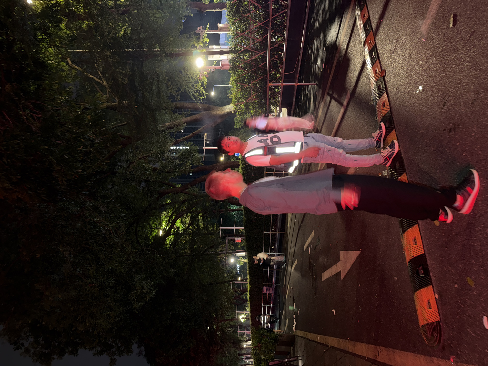 The image depicts two individuals standing on a pedestrian street in Tianhe District, China. A person wearing a reflective safety vest is standing beside another person wearing a grey shirt and black pants. The street is lined with trees and a metal fence with hedges behind it. There are streetlights illuminating the area. The street has markings, including a white arrow and yellow and black speed bumps.