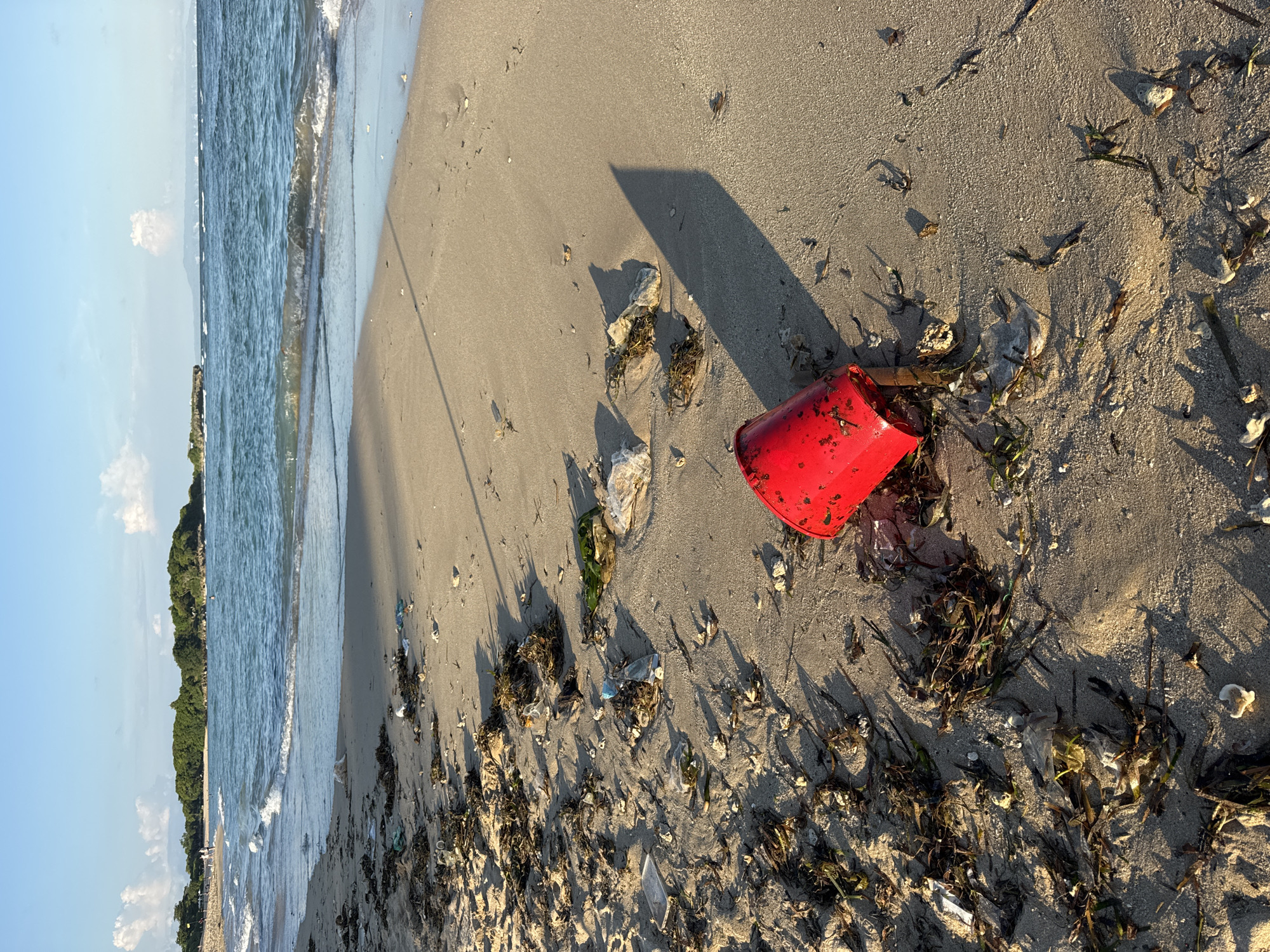 A low-angle, medium shot shows a sandy beach, the ocean, and the coast in Unknown, Indonesia. A red plastic bucket is lying on its side on the sand to the right of the image center. There is seaweed and other marine debris scattered on the sand around the bucket. The sand is damp and appears to be packed in some areas and loose in others. The ocean is visible in the background, with small waves rolling in toward the shore. The coast is to the left of the image and features a line of trees. The sky is partly cloudy.
