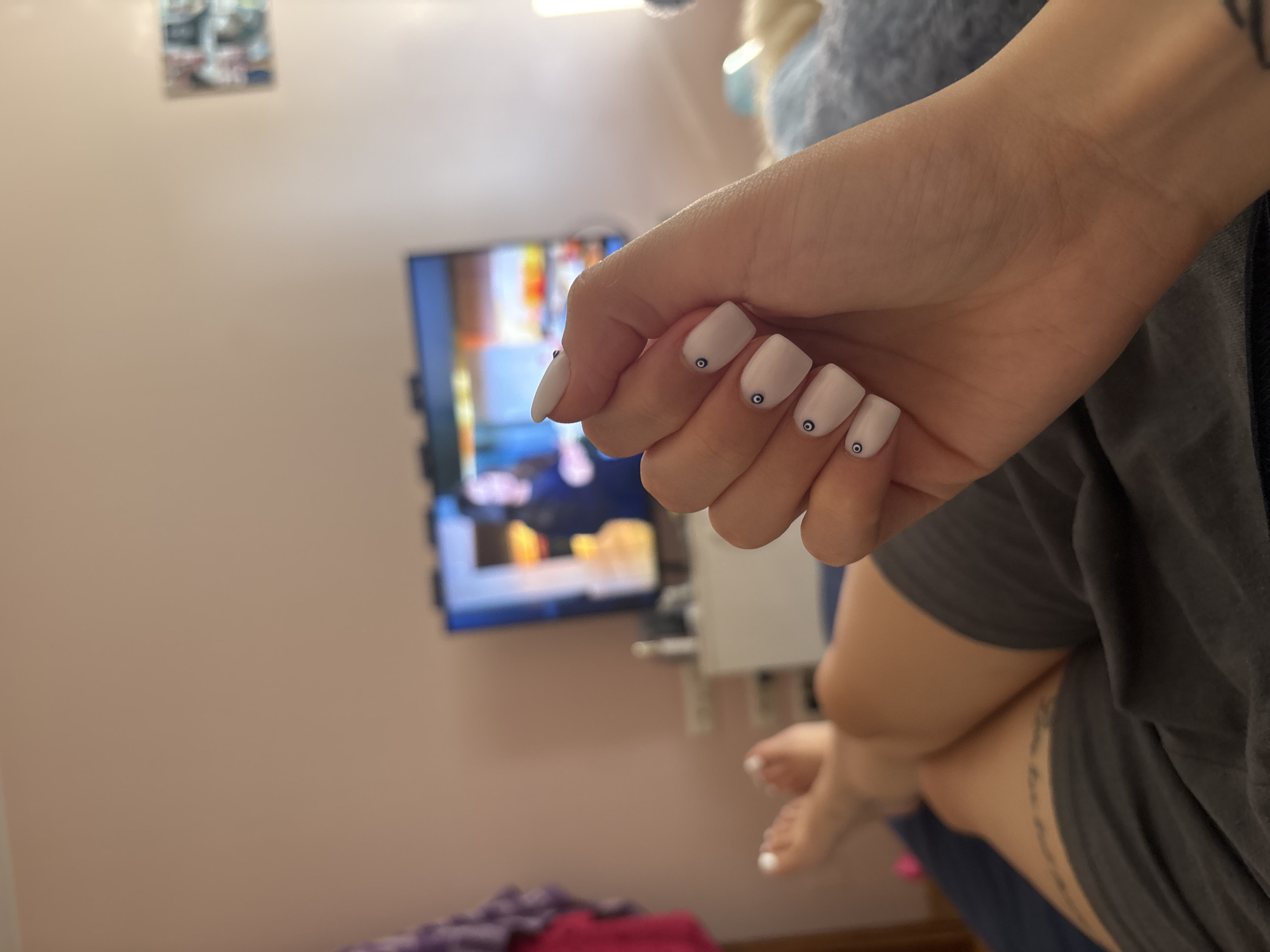 A medium shot captures a human hand with white-painted nails, each adorned with a small, dark blue "evil eye" design, positioned in front of a blurred background. The blurred background reveals a television screen showing a vibrant, colorful scene, a wall-mounted media console, and a lilac-colored wall. The lower part of the frame shows a glimpse of a person's legs and feet, with painted white toenails. The person is wearing a dark grey garment, and a tattoo is visible on their thigh. The location is suspected to be an interior space in Sofia, Bulgaria.