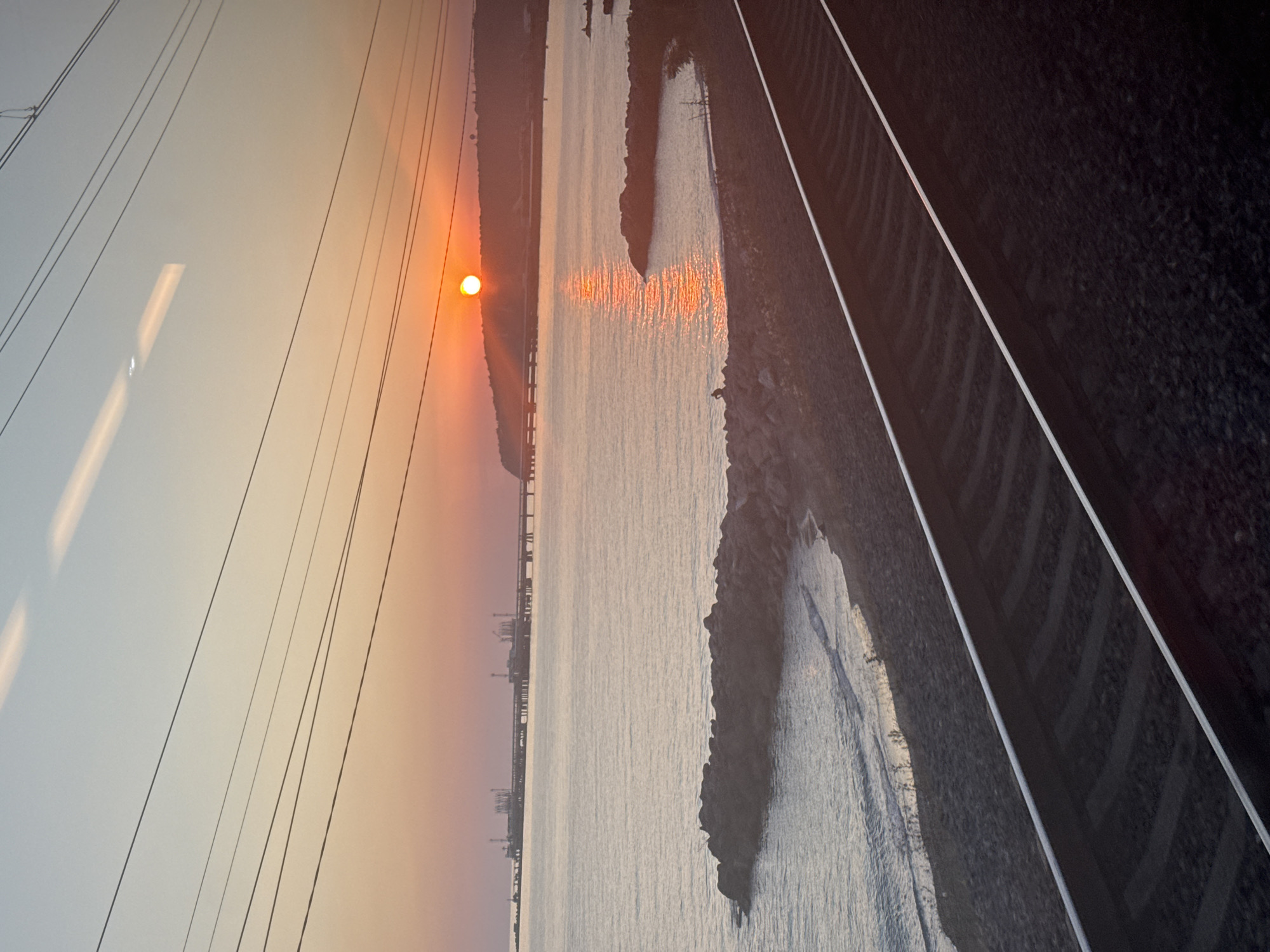 An exterior, medium shot captures a sunset over the Black Sea from the perspective of a moving train, likely near Tuapse, Russia. The sun, a bright orange orb, sits on the horizon, casting a shimmering reflection across the calm water. The sky transitions from a soft orange near the sun to a pale blue at the top, with several overhead wires crisscrossing the frame.
In the distance, a long industrial pier juts out into the sea, with structures and cranes visible. The shoreline is rocky, with a small beach area in the foreground. Two sets of train tracks dominate the bottom half of the image, stretching into the distance. A single, dark silhouetted figure is seated on the rocks near the water's edge. The scene is peaceful and serene, with the warm hues of the sunset creating a tranquil atmosphere.