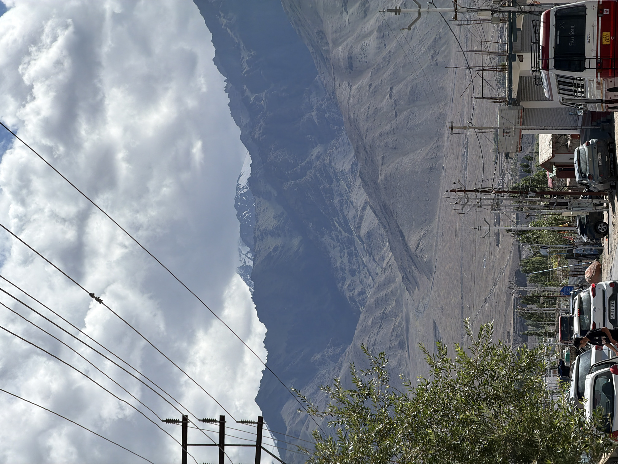 This image depicts a street scene in Leh, India, with a backdrop of imposing mountains. A cloudy sky with patches of blue overhead dominates the upper portion of the frame. Multiple power lines crisscross the sky and are supported by utility poles. In the foreground, a green leafy tree partially obscures the view of a street lined with parked cars, including a white Hyundai hatchback and a white minibus with red trim. A person, appearing to be a young male wearing a black t-shirt, walks on the street. The minibus has "Free Soul" written on its rear window. Beyond the street, the landscape transitions into a dry, barren terrain that slopes upwards towards rugged, dark-colored mountains. The snow-capped peaks of higher mountains are visible in the distance. Buildings and smaller structures are scattered along the roadside and further into the distance, suggesting a settlement amidst the mountainous terrain. The overall impression is one of a functional street in a mountainous, high-altitude region.